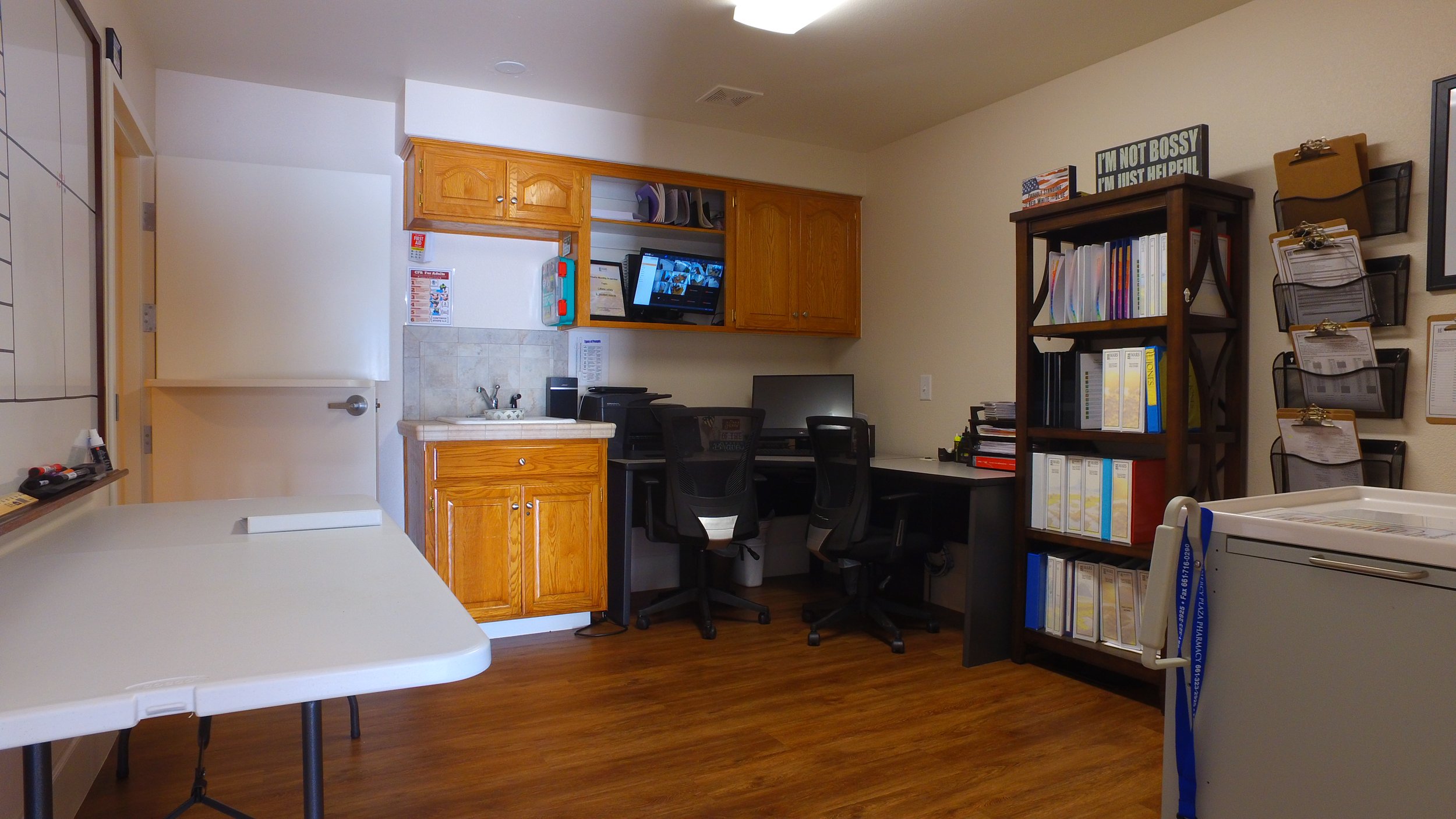 Office room with wood flooring, a whiteboard, a table, a small kitchenette with wooden cabinets, a computer desk with two black chairs, a monitor, a clipboard, and bookshelves with binders. Signage and wall-mounted files are also visible.