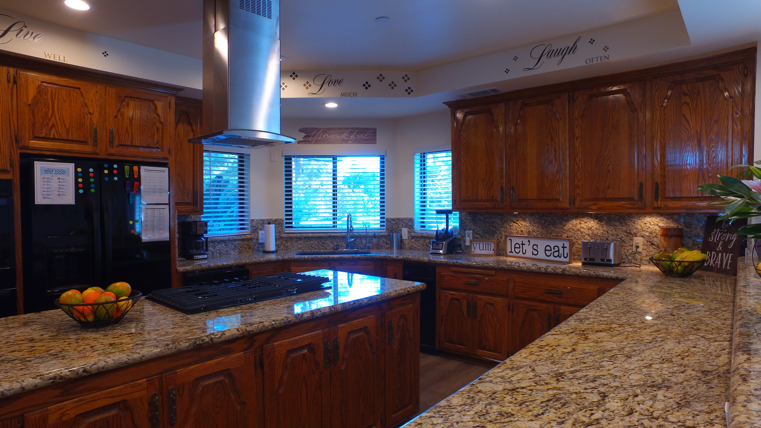 Kitchen with wooden cabinets, granite countertops, a black refrigerator with colorful magnets, a stovetop, and a range hood. There are windows with blinds, decorative wall words, and various small appliances on the counter, including a blender and to