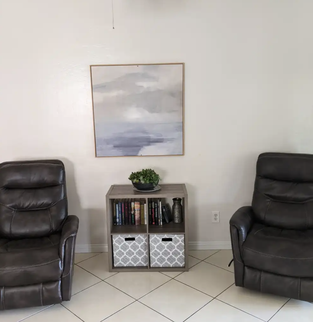 Living room with two brown leather chairs, a small bookshelf with books and decorative storage bins, a plant on top, a framed abstract painting hanging on the wall, and tiled flooring.