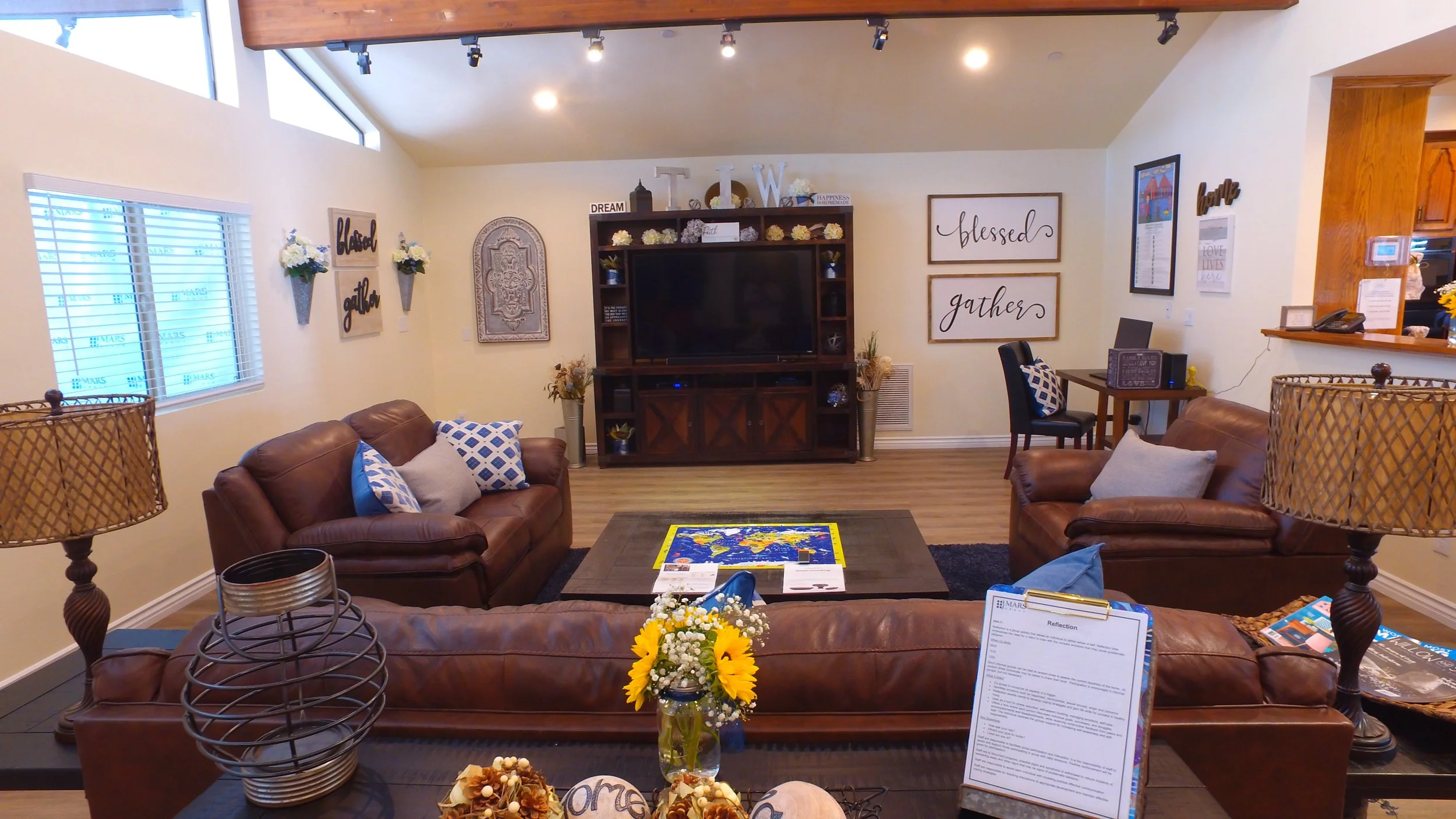 Living room with two brown leather sofas, a dark wood coffee table with a world map, vase of sunflowers, and various decorative items. A large television is centered on a dark wood entertainment center with shelves. The walls are decorated with frame