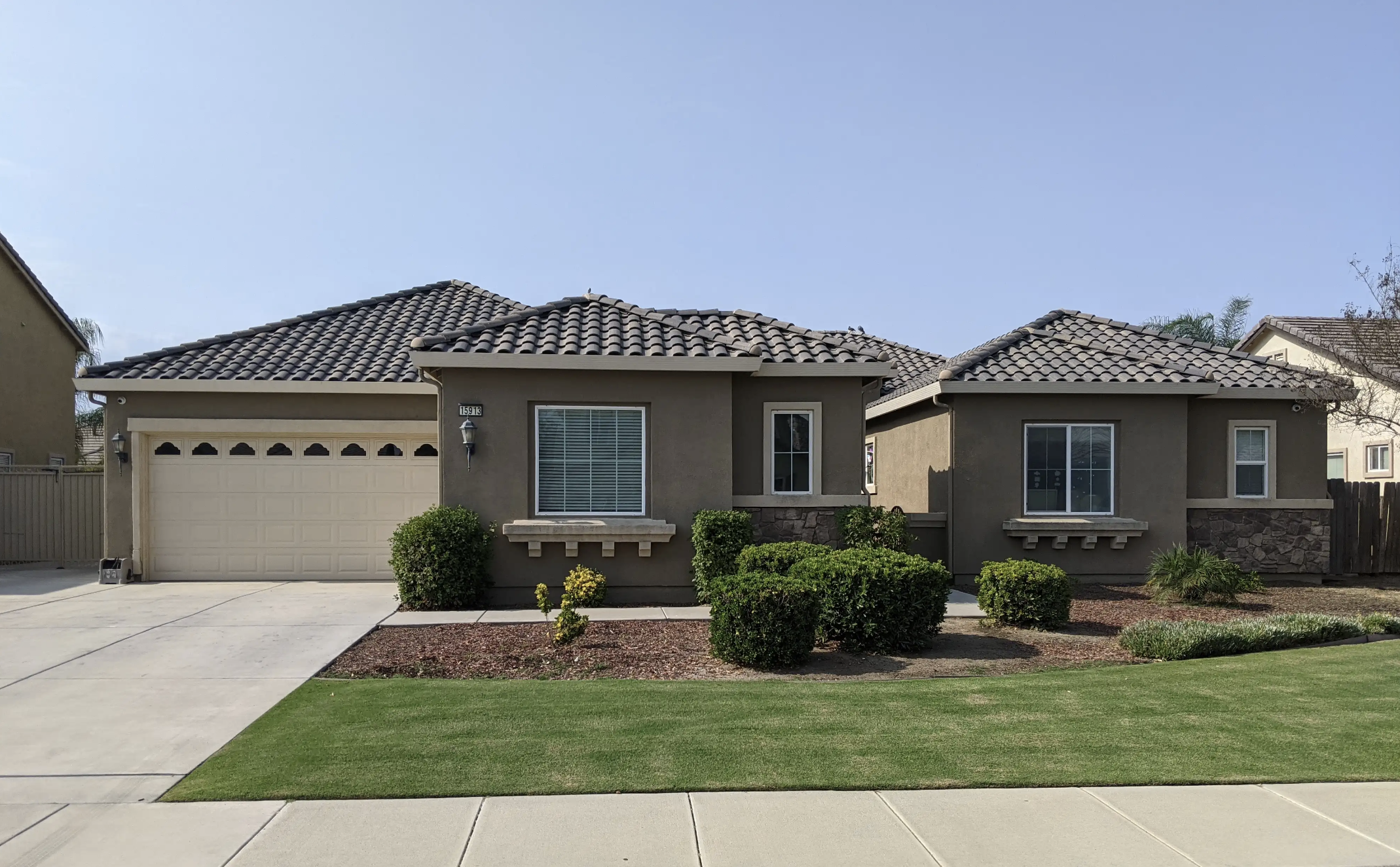 Front view of a single-story house with brown exterior, gray tiled roof, two large windows, a garage with a white door, and well-maintained green lawn and shrubs.