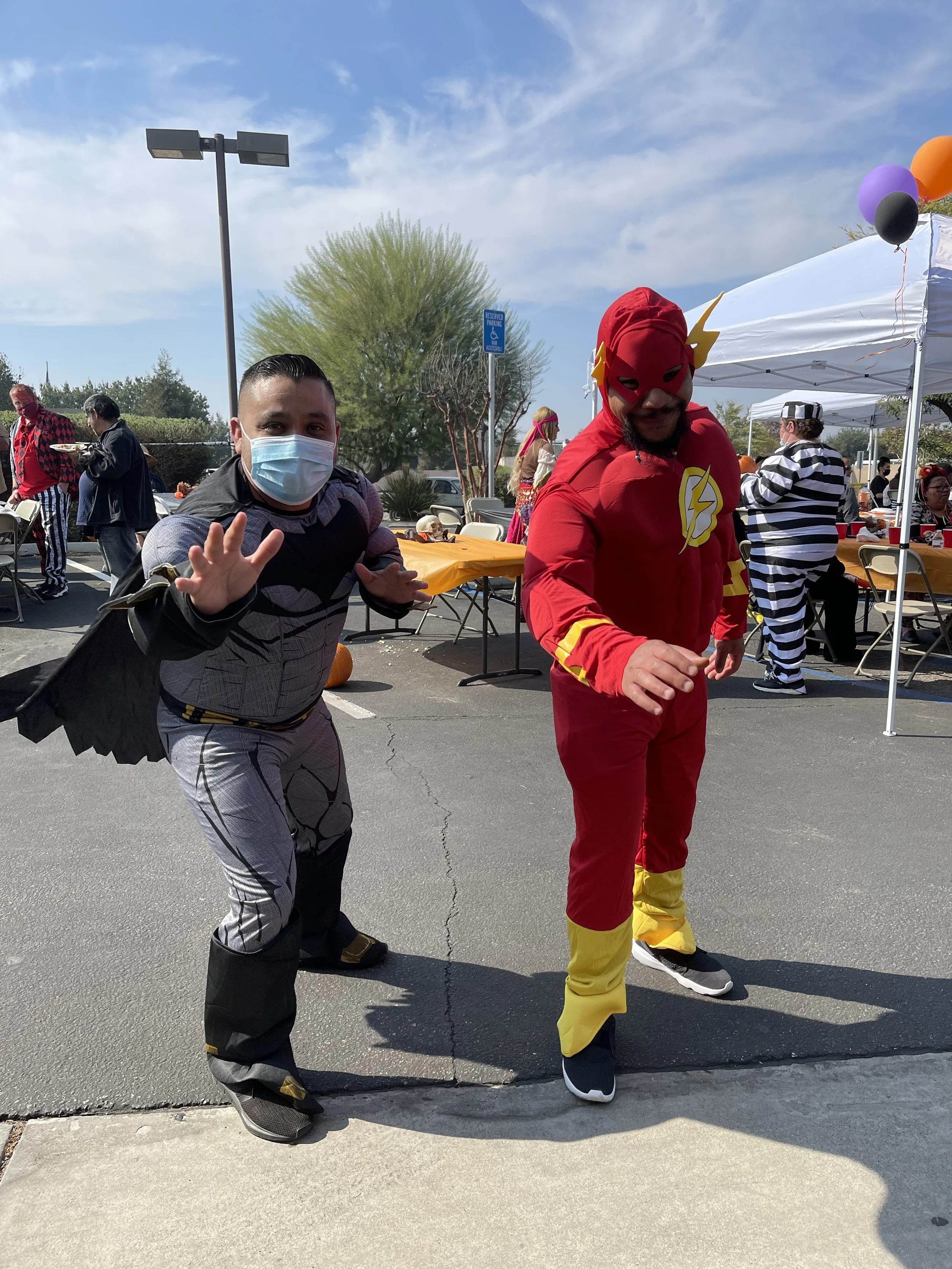 People dressed in costumes, one as The Flash and one as Black Panther, at a costume party outdoors with tables, chairs, and balloons.