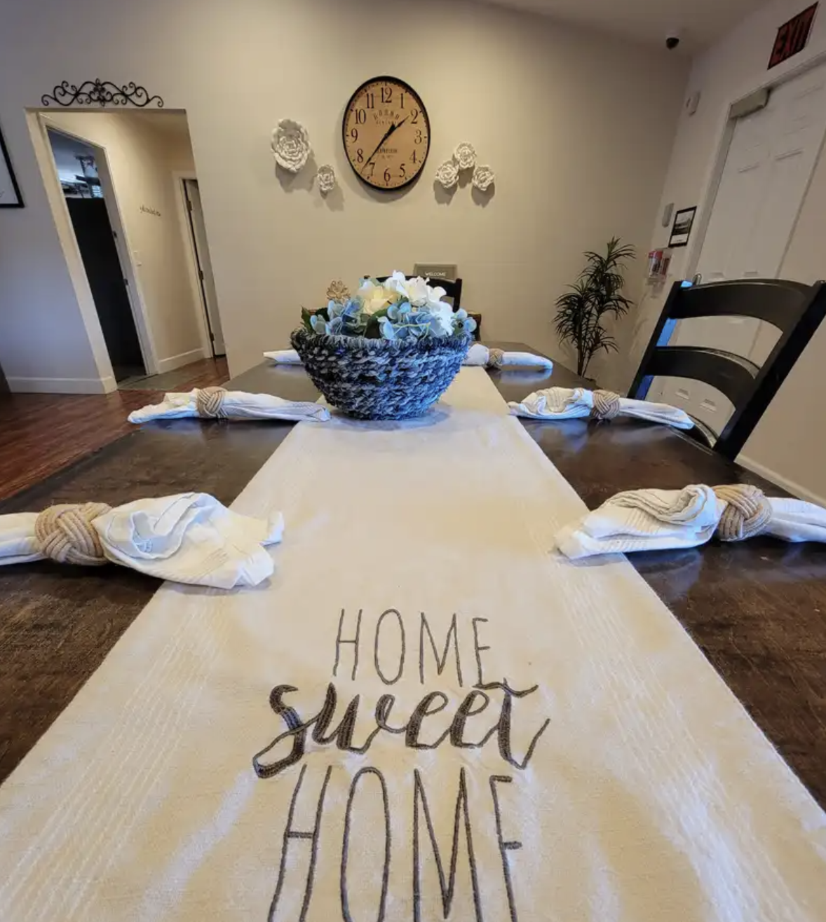 Decorated dining table with a beige table runner that has the words "Home sweet home" written on it. The table has a basket of white and blue flowers as a centerpiece and is set with white napkins tied with beige knotted napkin rings. In the backgrou