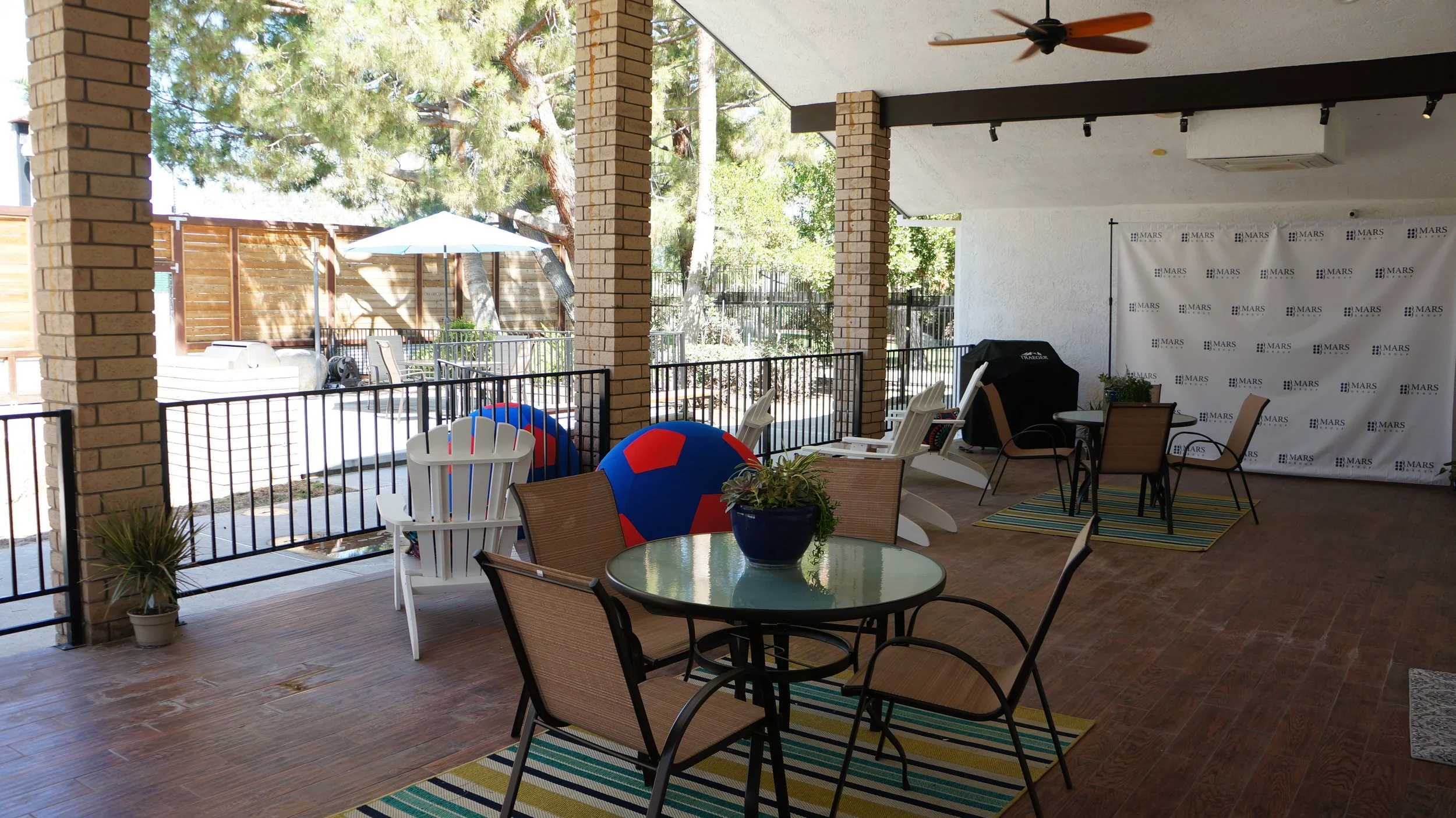 Covered patio area with outdoor furniture, including a round table with a plant, chairs, lounge chairs, and large colorful beach balls. The patio has a wooden floor, brick columns, and a railing, with trees and a grill outside. There is a backdrop wi