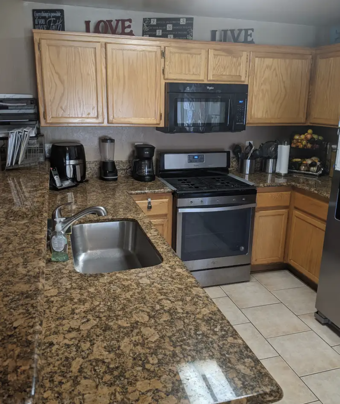 Kitchen with wood cabinets, granite countertops, stainless steel stove and microwave, coffee maker, blender, paper towels, fruit basket, and a sink with soap.