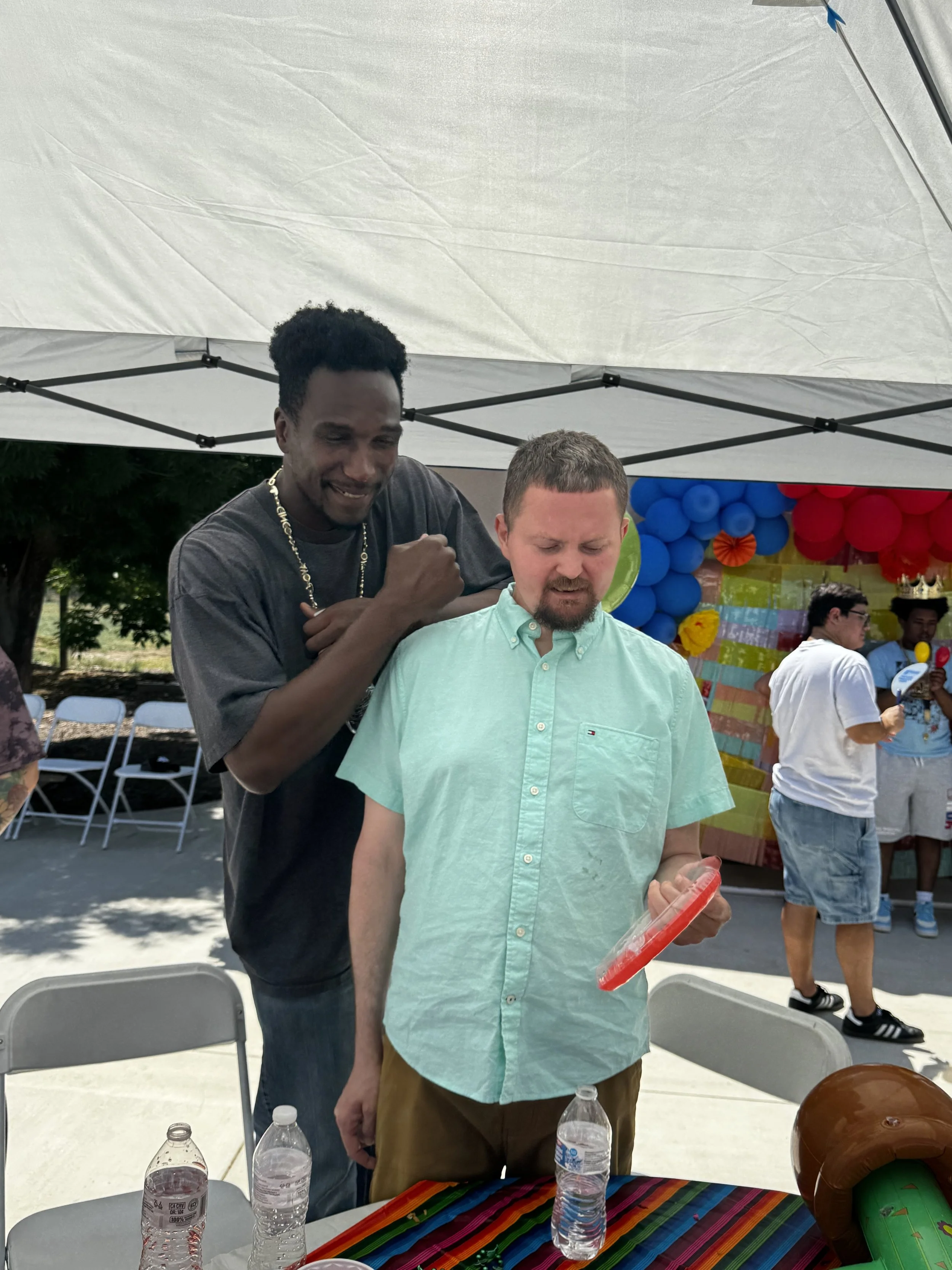 Two men at a colorful outdoor celebration, one with dark skin and black hair smiling and giving a fist bump, the other with light skin and brown hair looking at an object in his hand, under a white tent with balloons and a colorful backdrop in the ba