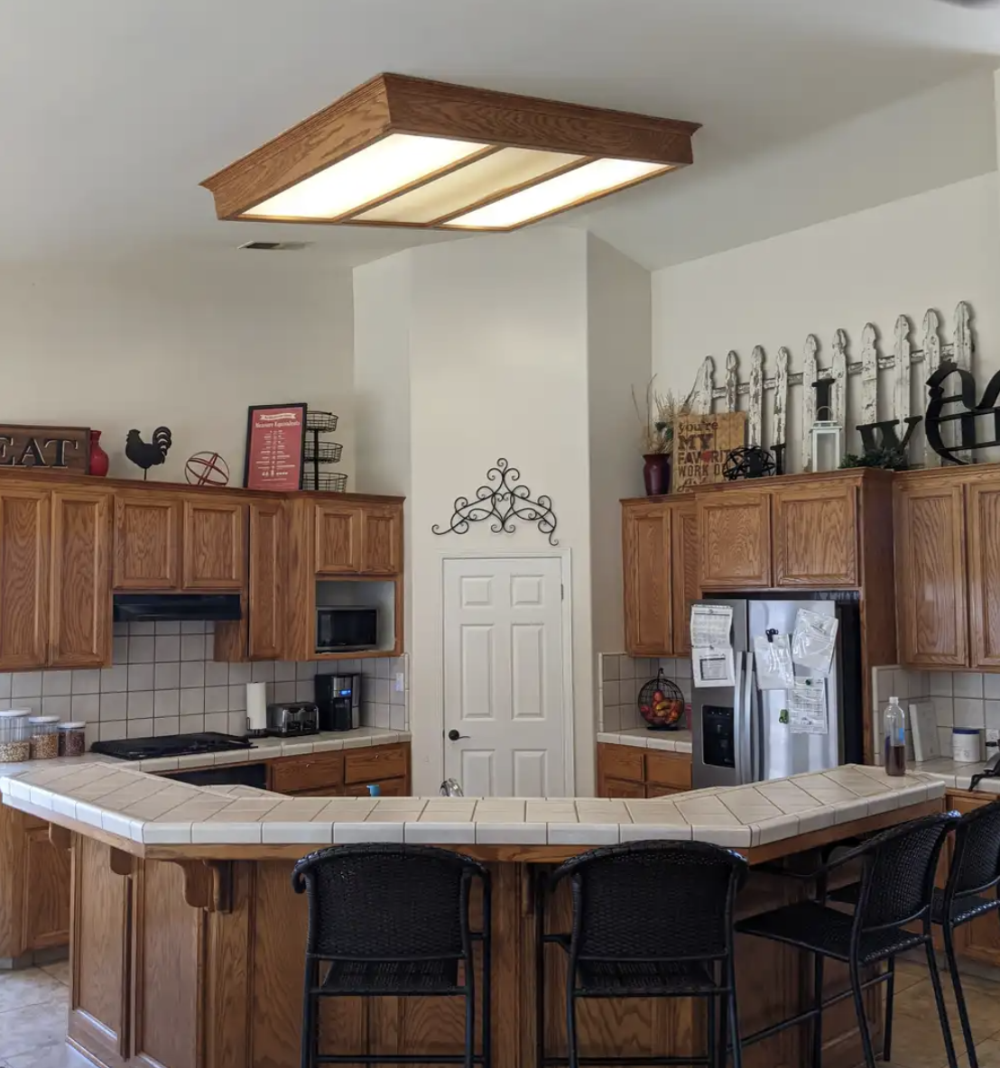 Kitchen with wooden cabinets, beige tiled countertops, a white door, and decorative items on top of the cabinets, including a rooster figurine and a red vase. There is a stainless steel refrigerator, a microwave, and a coffee maker.