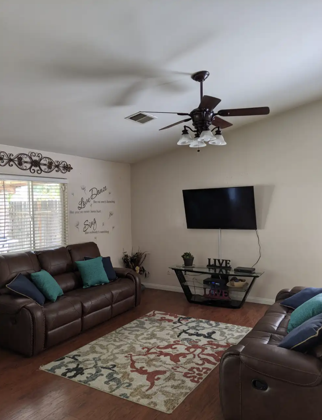 A living room with two brown leather sofas, a patterned area rug, a wall-mounted TV, a glass TV stand with decor items, a ceiling fan, a window with blinds, and wall decals with inspirational quotes.