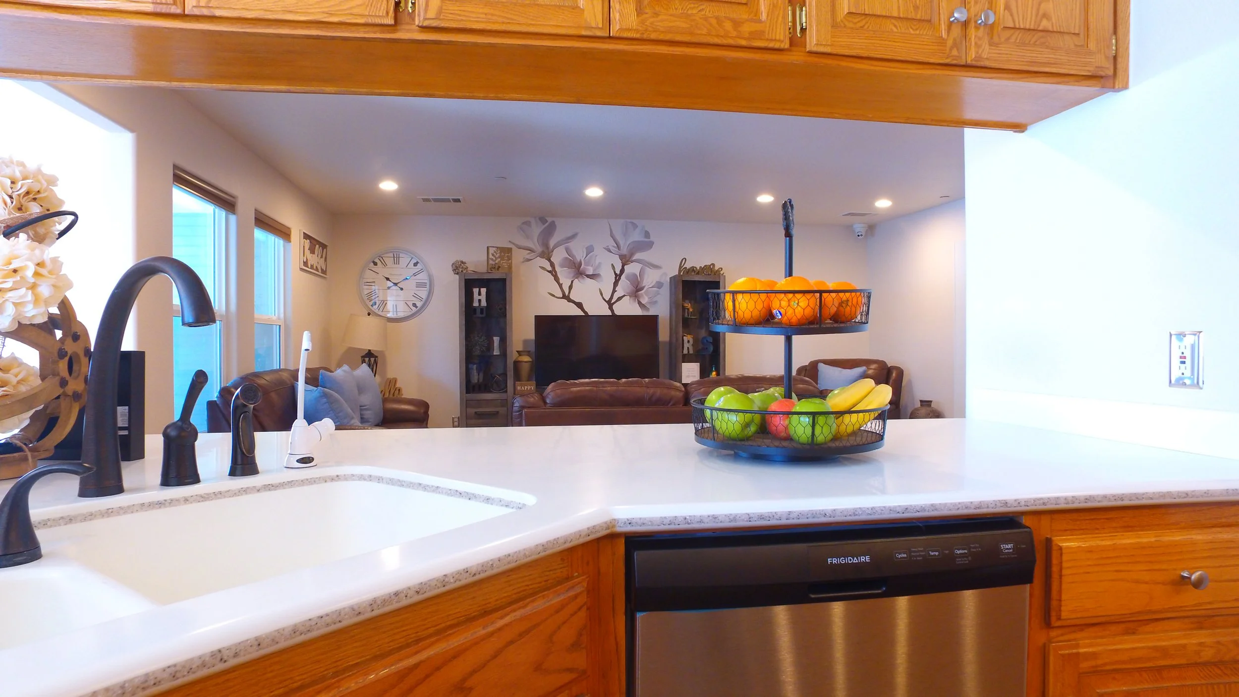 Kitchen counter with a fruit bowl containing bananas, apples, and oranges, overlooking a living room with leather sofas, a TV, and decorative wall art.