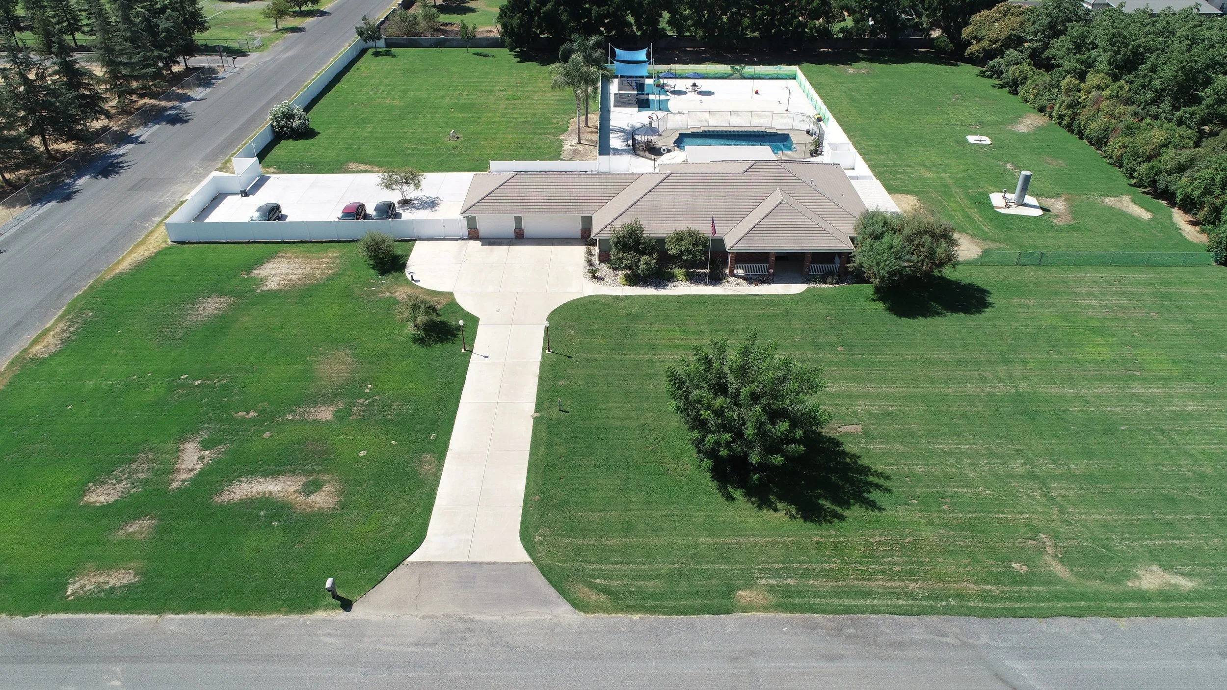 An aerial view of a property with a house, a driveway, a fenced-in playground with a pool, parking area, and large grassy fields.