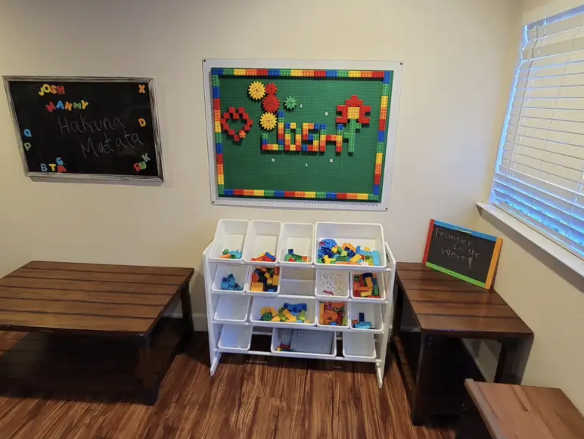 Play area with a lego wall mosaic spelling 'Liyana' beside a white lego bin shelf filled with various colorful lego pieces. There are two small wooden benches on either side of the lego shelf, a blackboard with the text "Hakuna Matata" and the names 