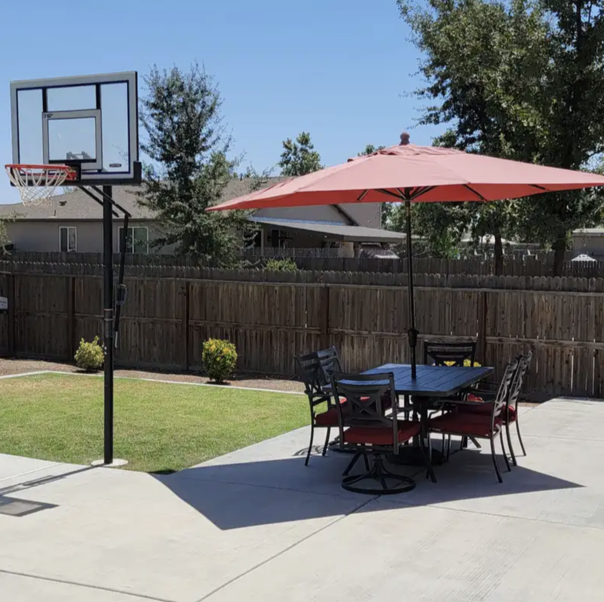 Backyard with a basketball hoop, a patio table with chairs and a pink umbrella, fenced yard, trees in background, clear sky.