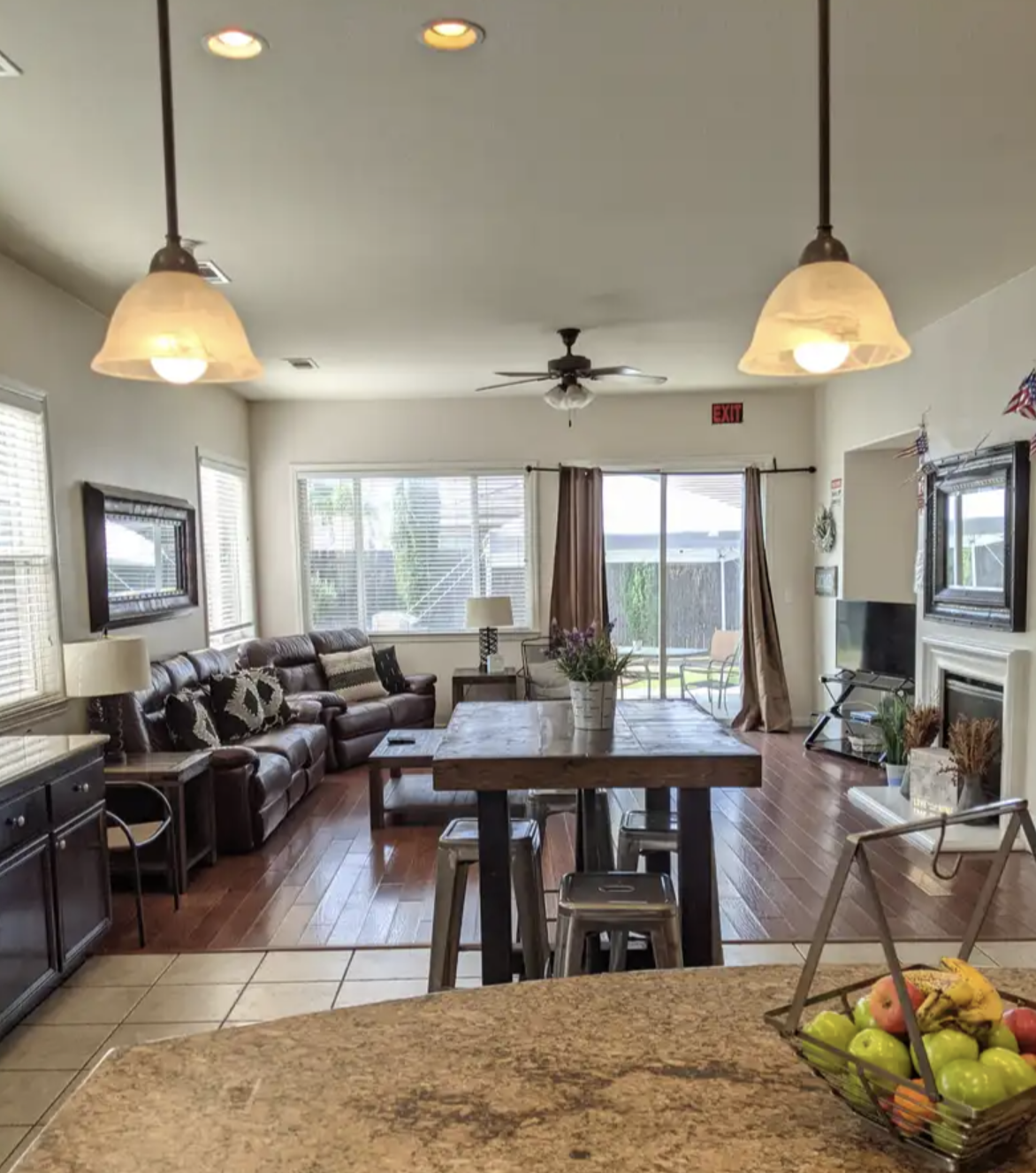 View of a living room with large windows, a brown leather sofa, a wooden dining table, a fireplace, a TV, and sliding glass doors leading outside. A bowl of fruit is on the counter in the foreground.
