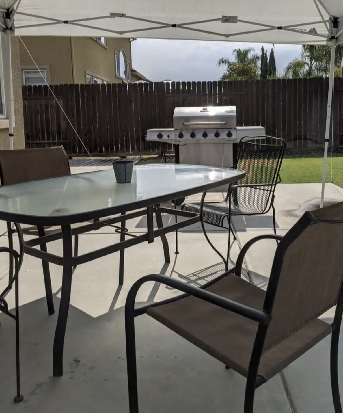 Outdoor patio area with a grill, a glass table with chairs, and a canopy overhead, enclosed by a wooden fence.