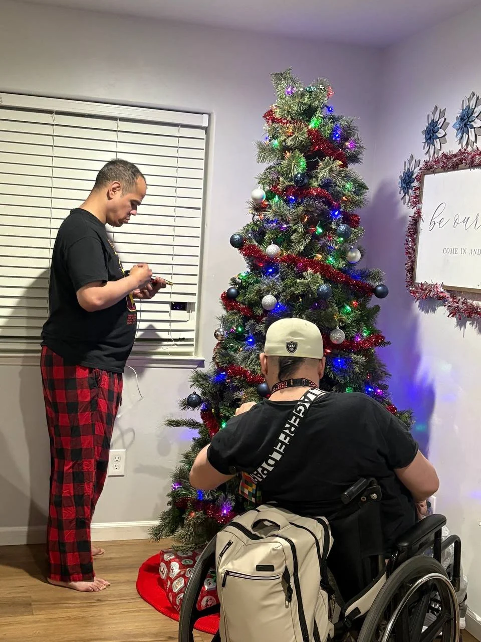Two young men are decorating a Christmas tree indoors. One is standing in plaid pajama pants, and the other is in a wheelchair wearing a cap.