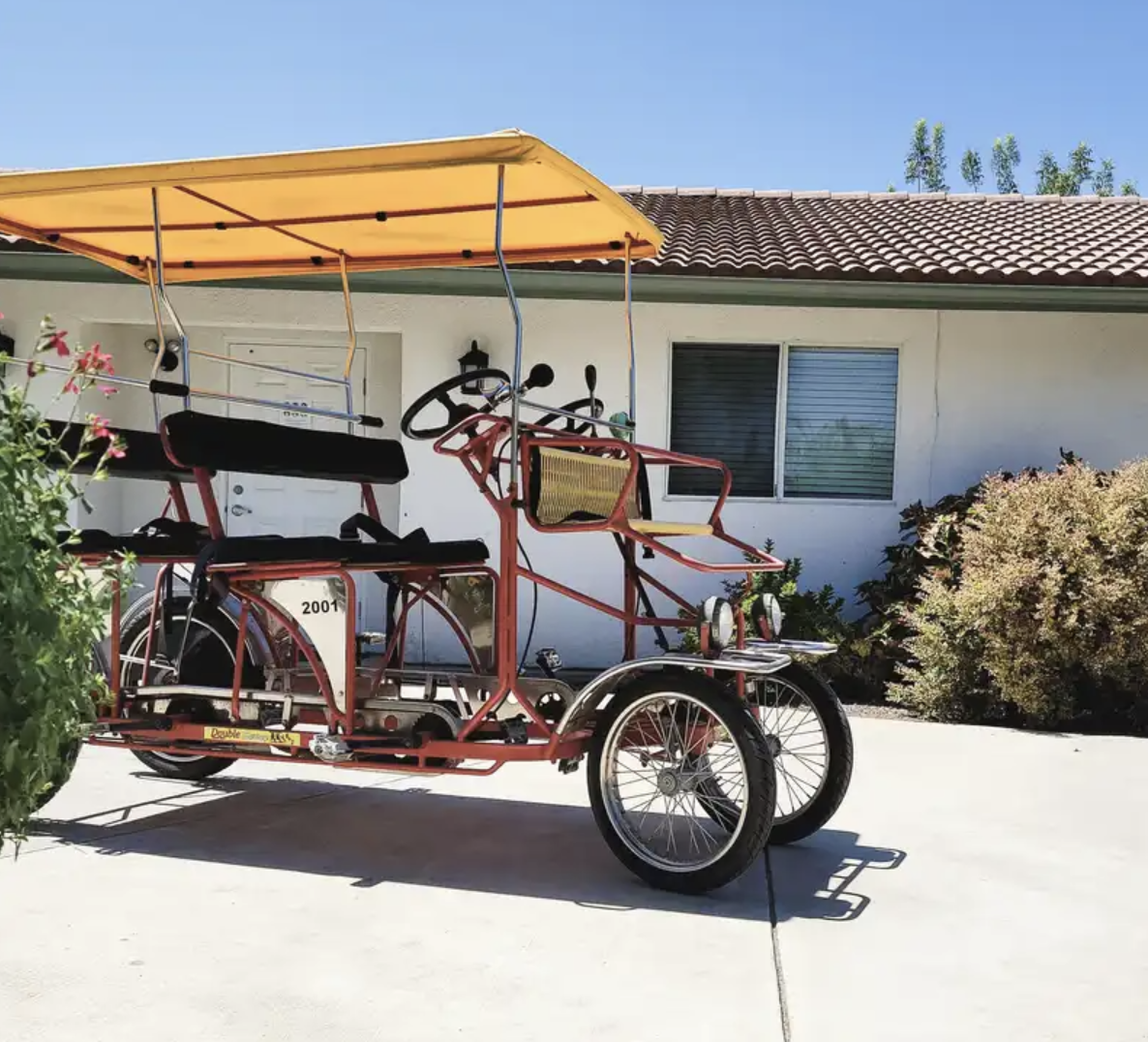 A red pedal-powered quadricycle with a yellow canopy, parked on a driveway in front of a white house with a window and greenery.
