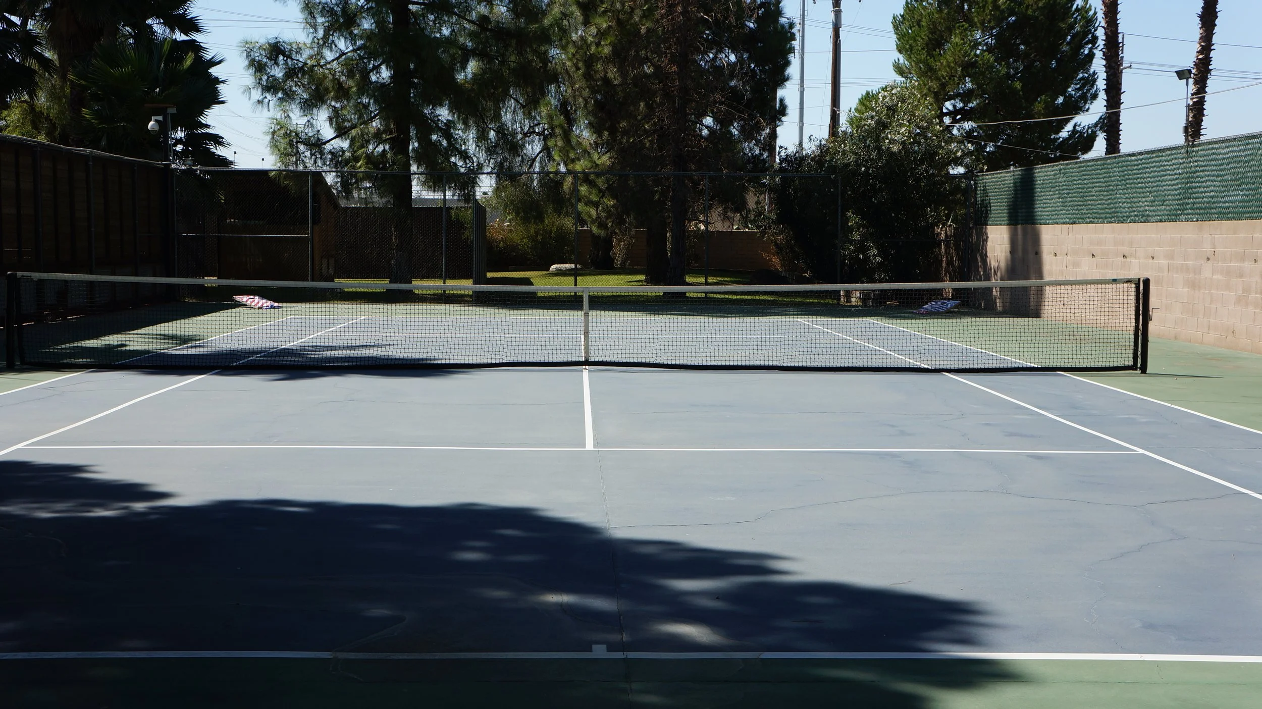 Empty outdoor tennis court with a net, surrounded by trees and a fence, and a brick wall on one side.