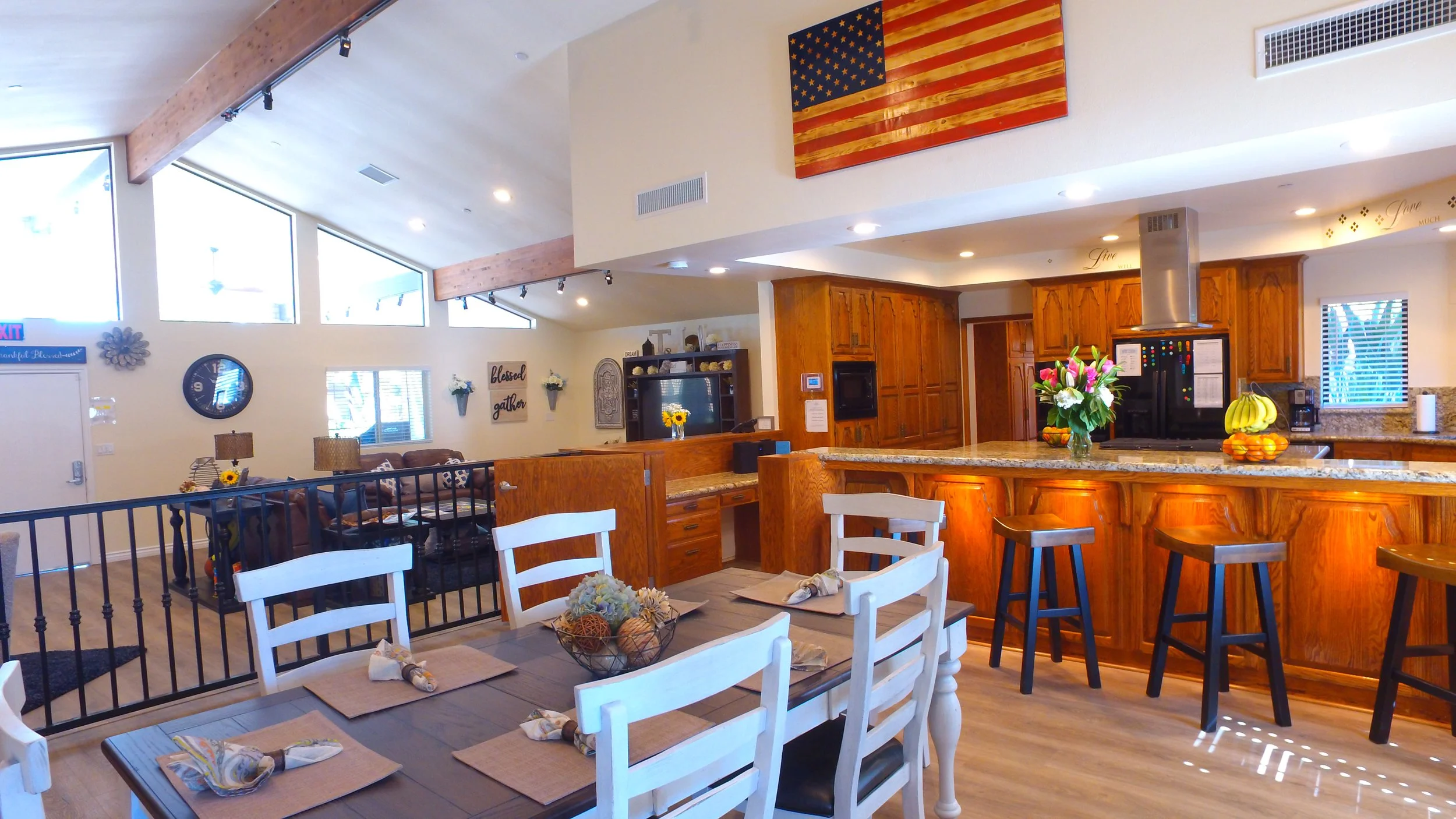 Open-concept kitchen and dining area with wooden cabinets, a granite island, and a dining table set with placemats and napkins, decorated with flowers in a bowl, in a cozy home interior.