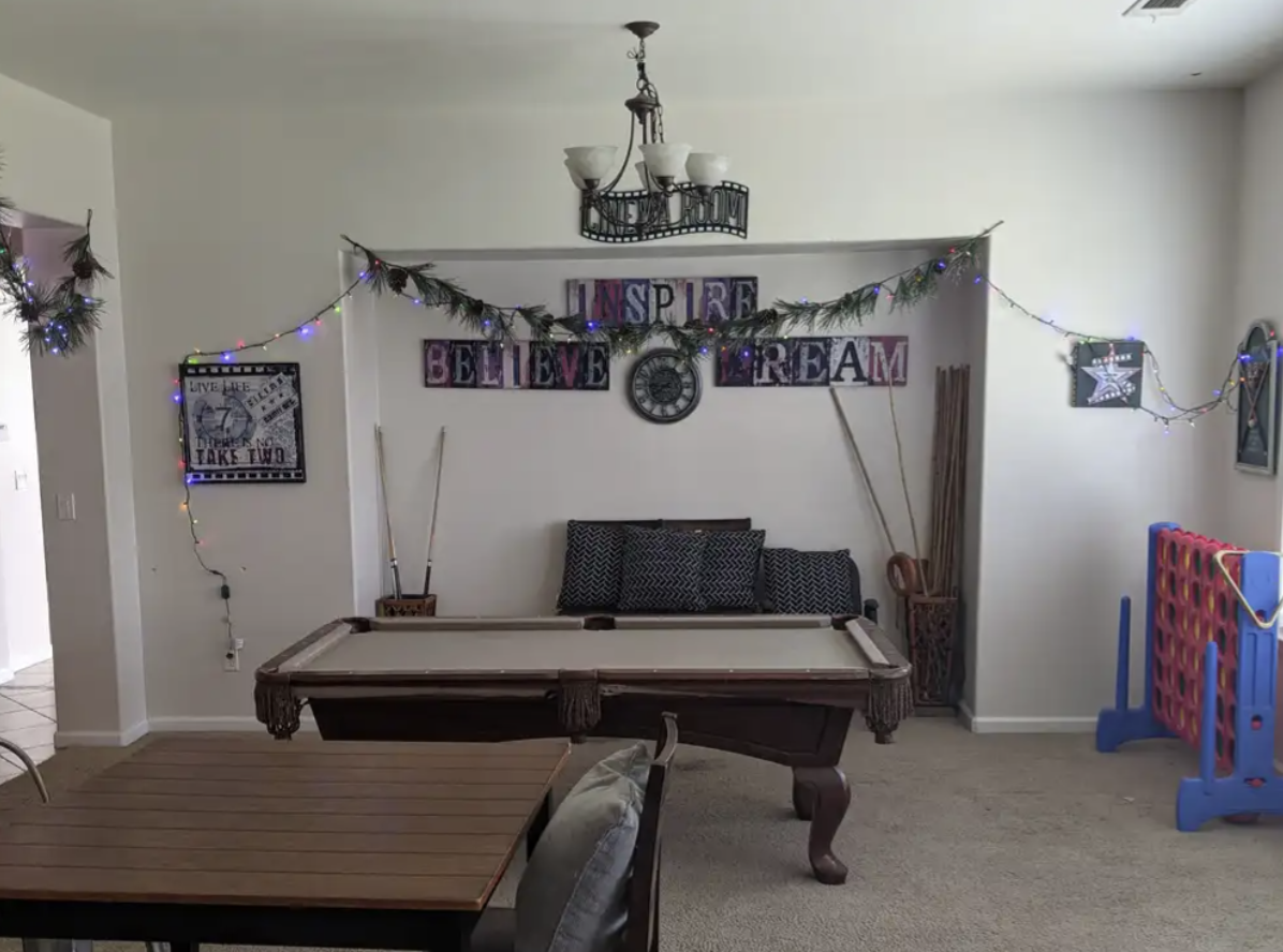 Living room decorated with string lights, evergreen garlands, and wall art. A pool table is in the center, with a couch and pillows against the back wall. There is a chandelier hanging from the ceiling, and some pool cues are standing in the corner. 
