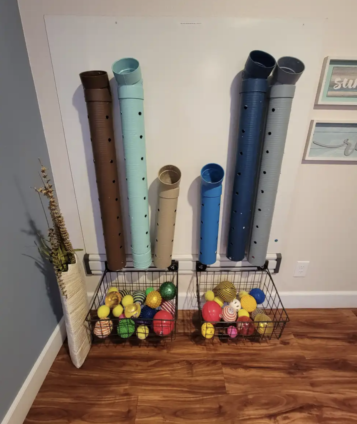 Colorful plastic pipes are mounted on the wall above two wire baskets filled with various colorful balls, in a room with wood flooring and walls decorated with framed artwork.