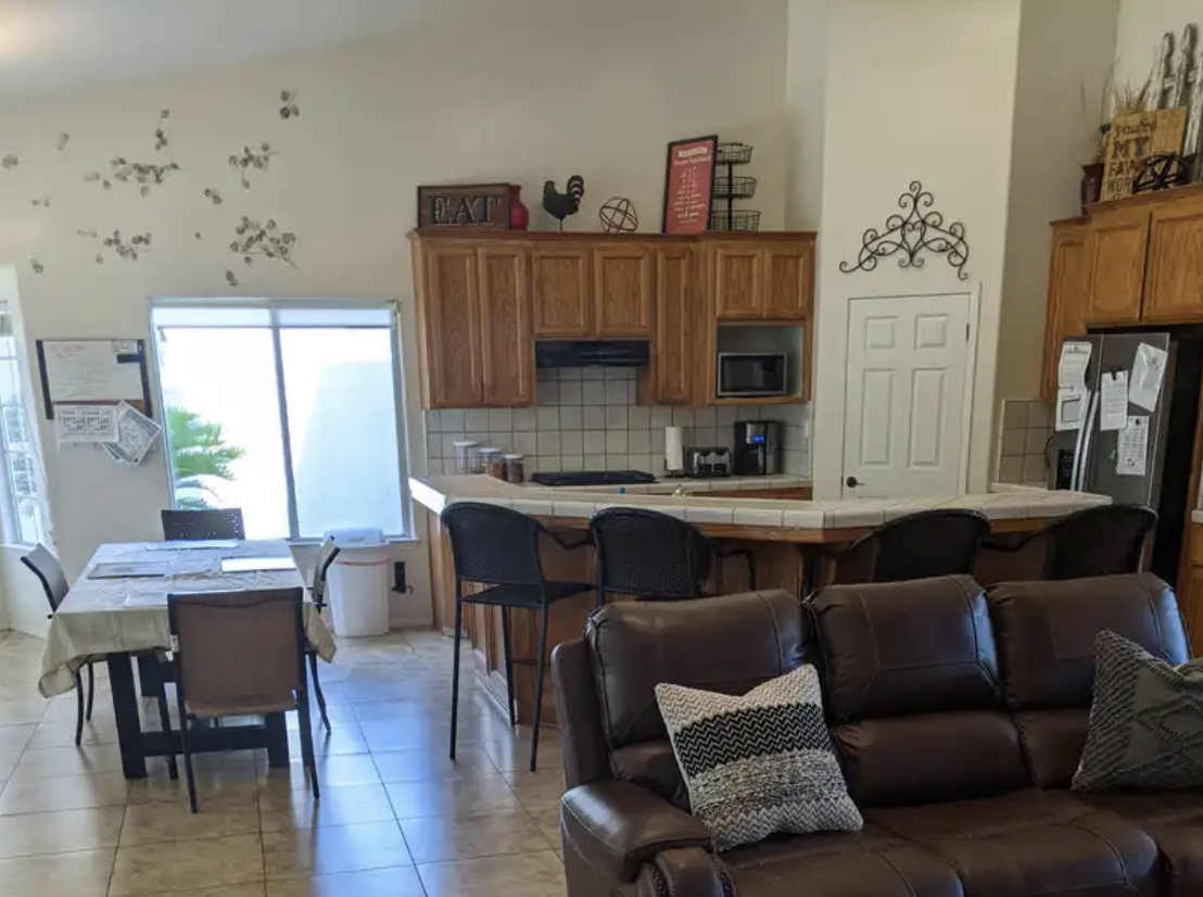 Kitchen and dining area with a sofa in the foreground. The kitchen has wooden cabinets, a refrigerator, microwave, and a breakfast bar with four chairs. There is a table with four chairs near a large window, and decorative items on top of the cabinet