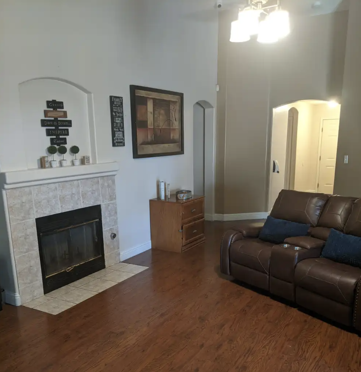 Living room with a fireplace, wooden floor, brown leather sofa with blue pillows, wall art, and a small wooden side table near the doorway.