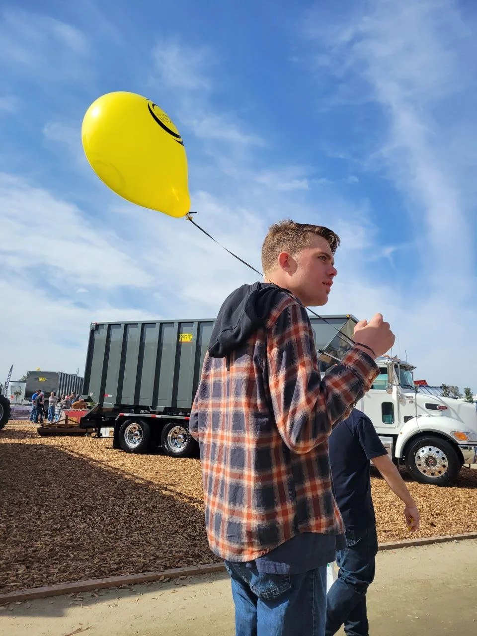 A person in a plaid jacket holds a yellow balloon with a large dump truck in the background.
