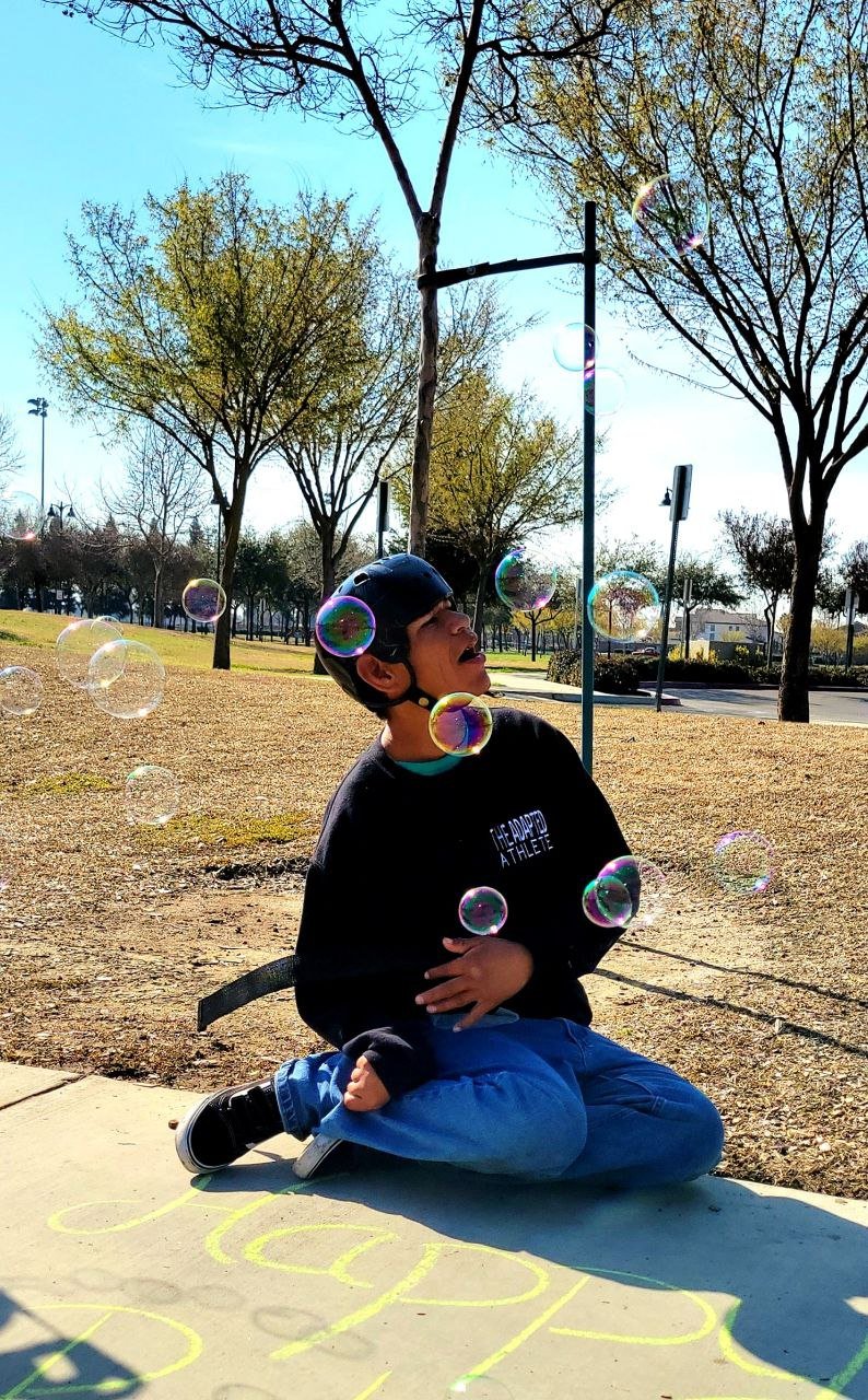 A person sitting on the sidewalk with the word 'Happy' written with chalk. There is a person sitting on the sidewalk with jeans, a black sweatshirt, and a safety helmet surrounded by rainbow bubbles.