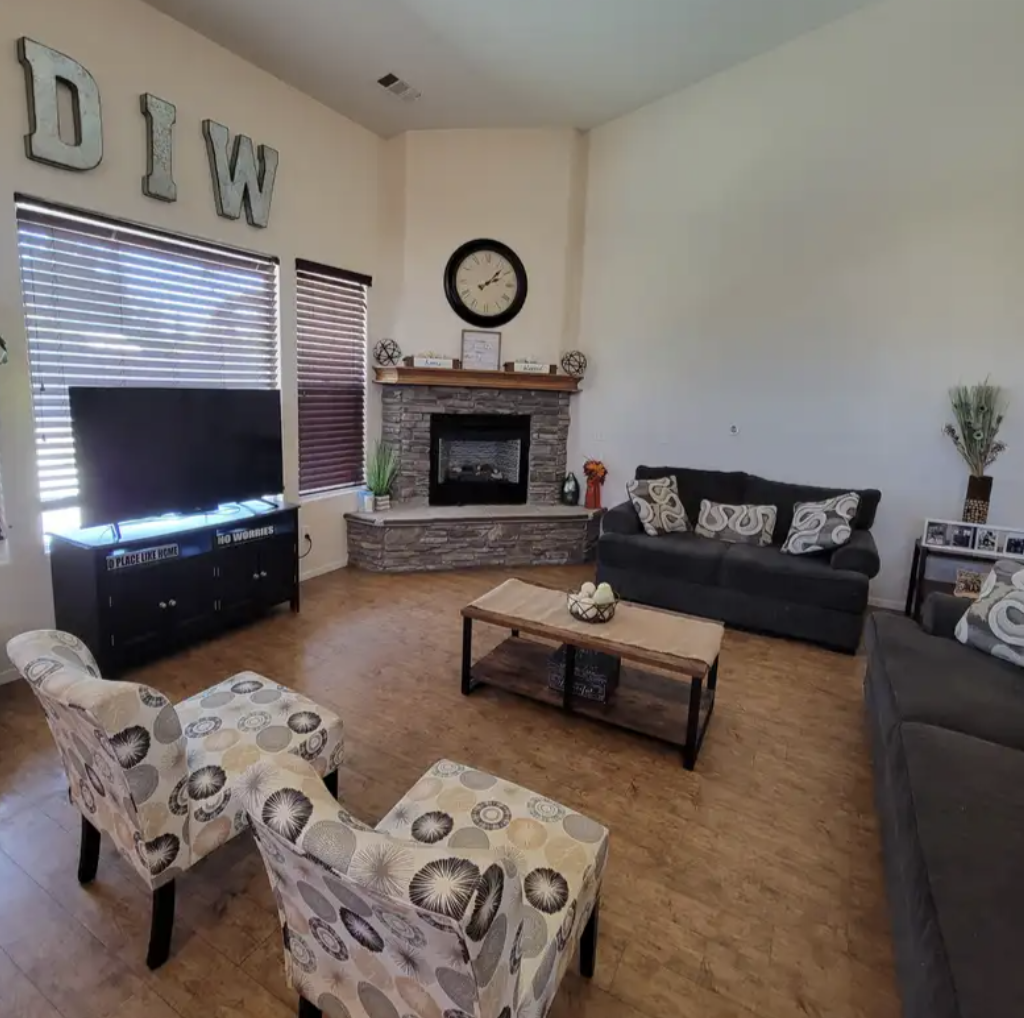Living room with a stone fireplace, a black TV on a stand, two patterned armchairs, two black sofas with decorative pillows, a wooden coffee table, and decorations on the mantel and walls.