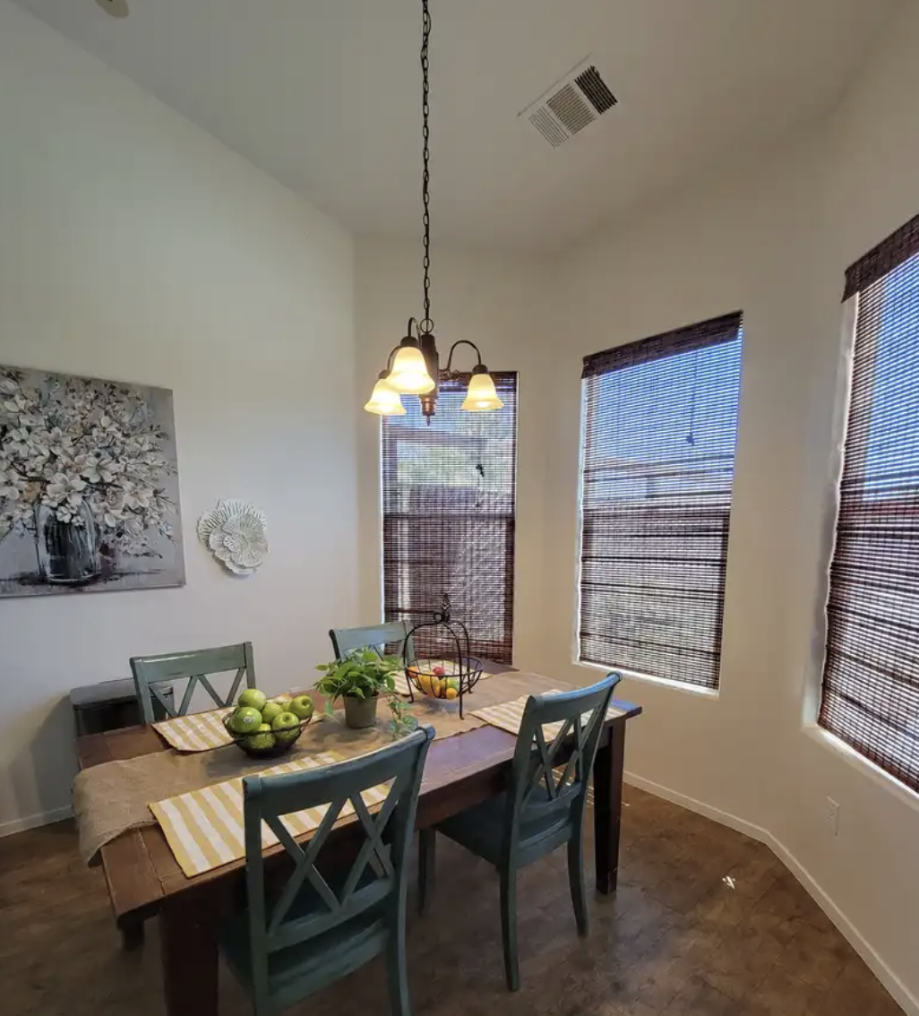 Dining room with wooden table, four blue chairs, green apples, potted plant, hanging light fixture with three lamps, windows with brown blinds, artwork on wall.