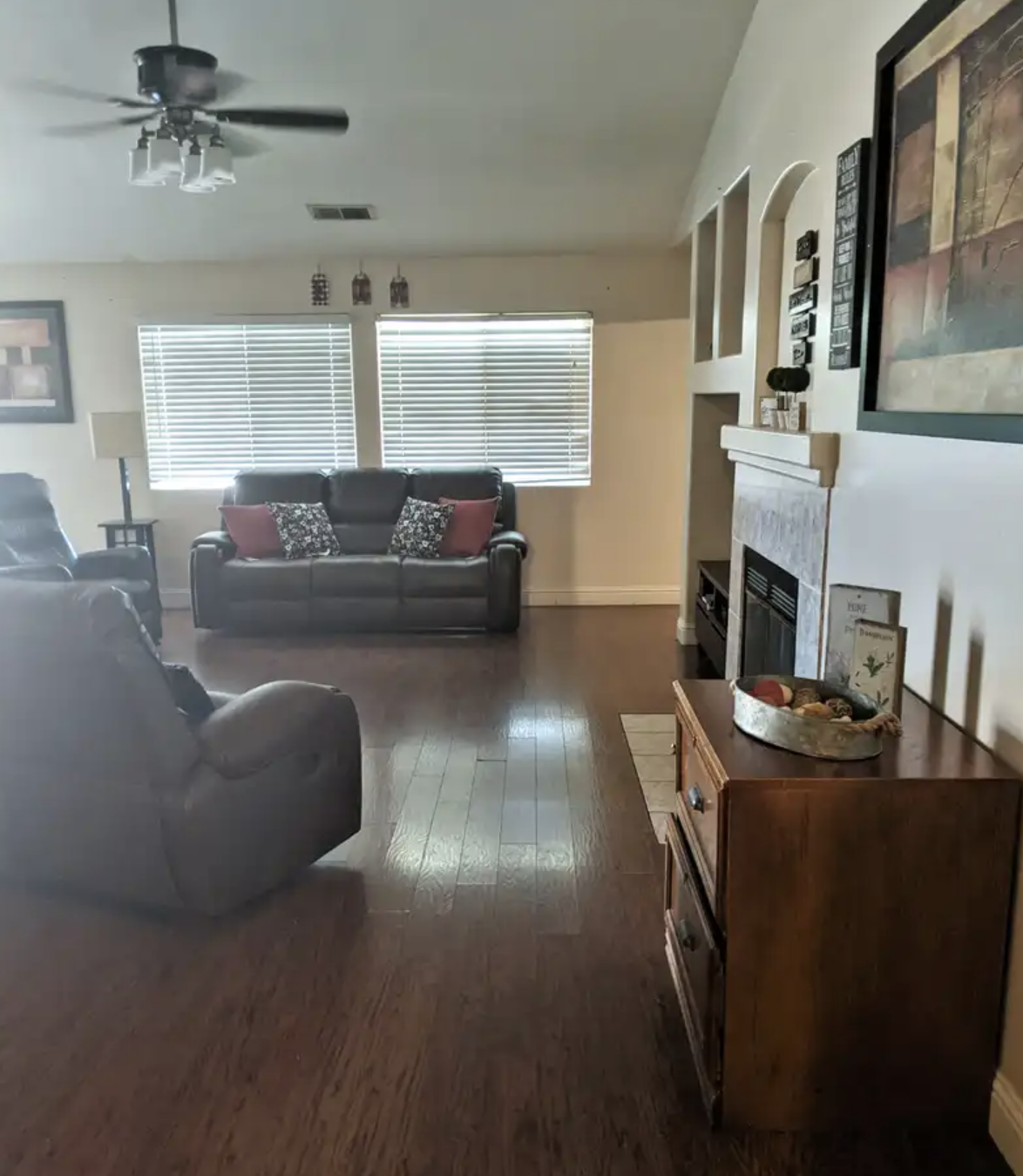 Living room with dark leather sofas, windows with horizontal blinds, a ceiling fan, a fireplace, a wooden console table, and wall art.