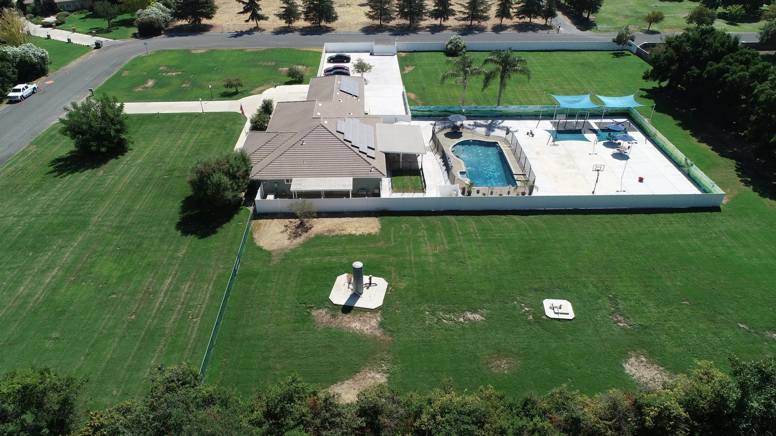Aerial view of a backyard with a pool, sports court, and surrounding green lawn and trees, enclosed by a white fence.