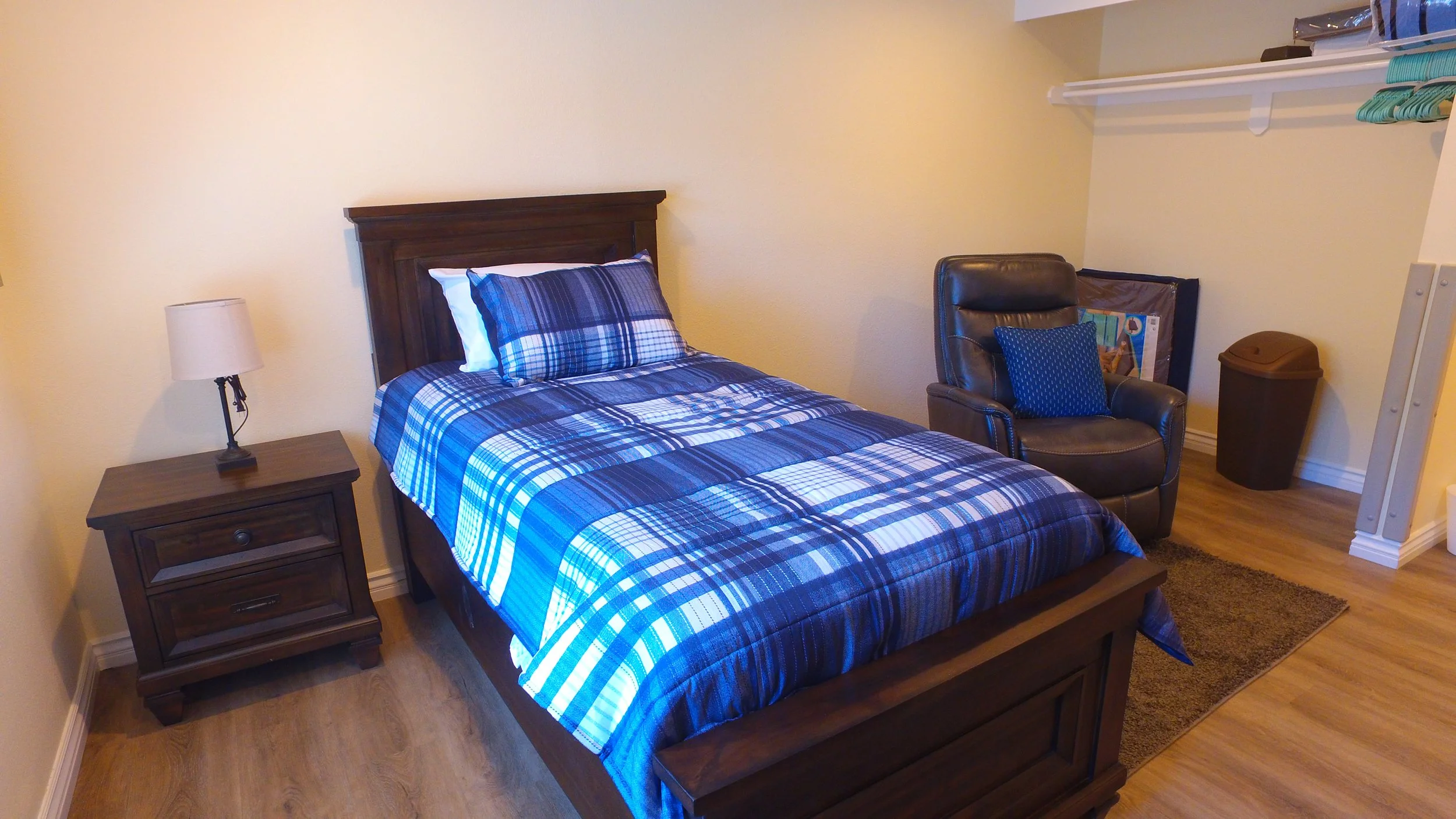 Bedroom with a wooden bed frame, blue plaid bedding, a nightstand with a lamp, a leather armchair with a blue throw pillow, a small rug, and an open closet with shelves and a trash can.
