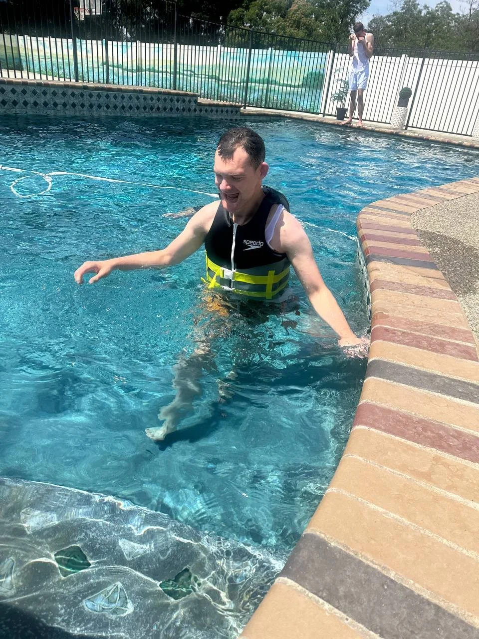 A man in a black life vest smiling and standing in a swimming pool, with a young person in the background near the pool's edge.