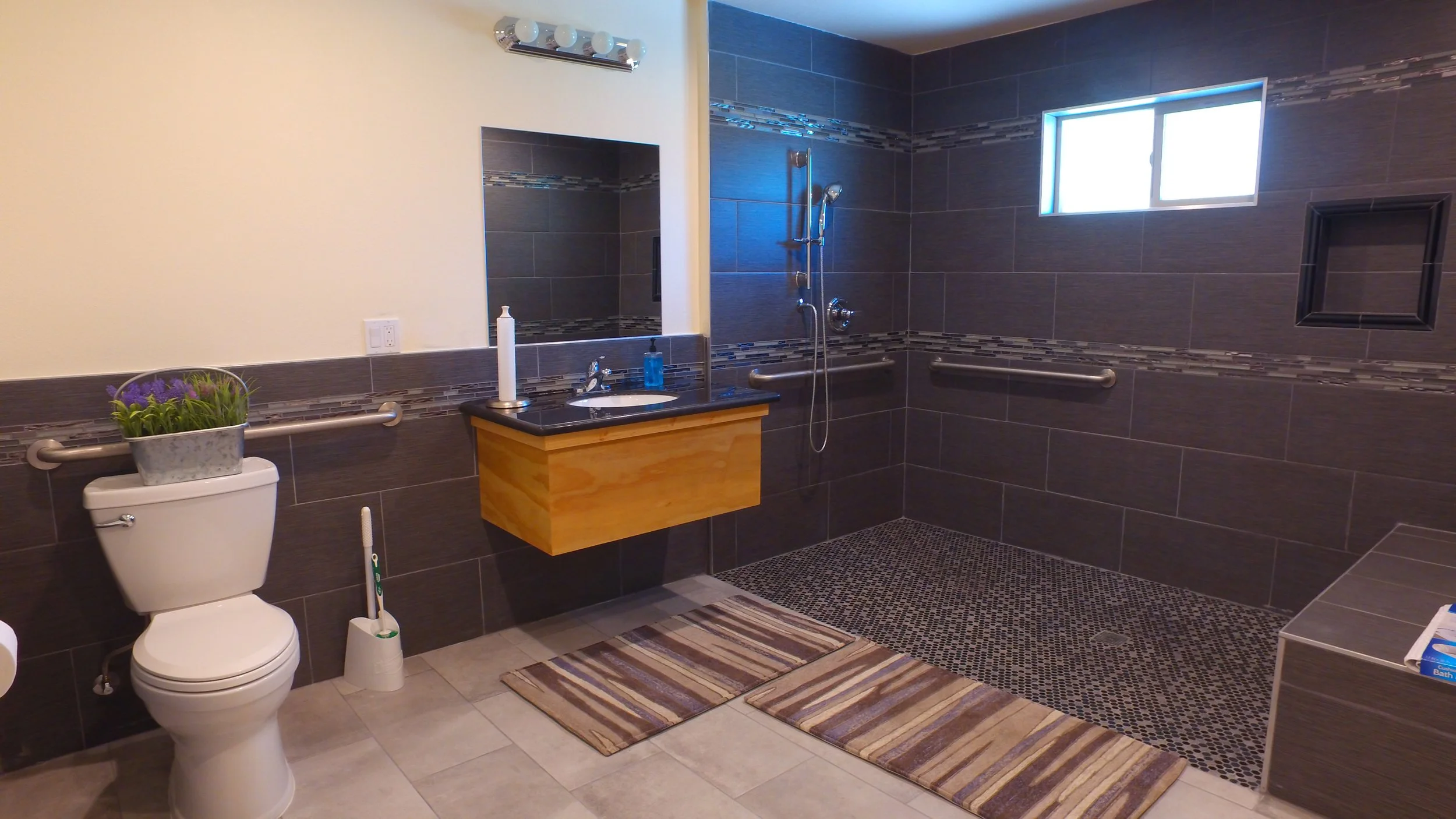 A spacious bathroom with a white toilet, a sink with a wooden cabinet, and a large walk-in shower with dark gray tiles, a small window, and safety grab bars.