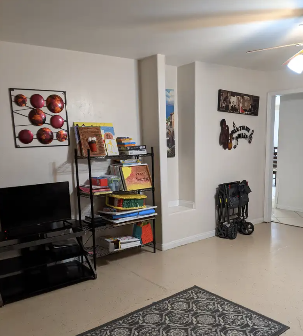 Living room corner with a black TV stand, a metal bookshelf with books and decorations, a wall decor with pink spheres, a small illustration of a coffee cup, and family photos on the wall. There's a folded stroller against the wall and a patterned ru
