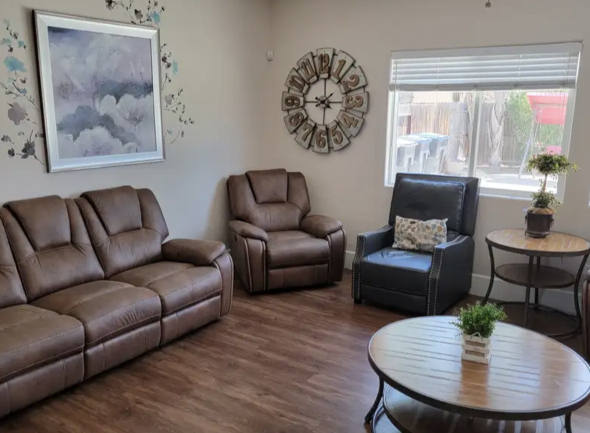 Living room with leather and fabric chairs, a wall clock, a window with blinds, a framed picture of clouds, and small tables with plants.