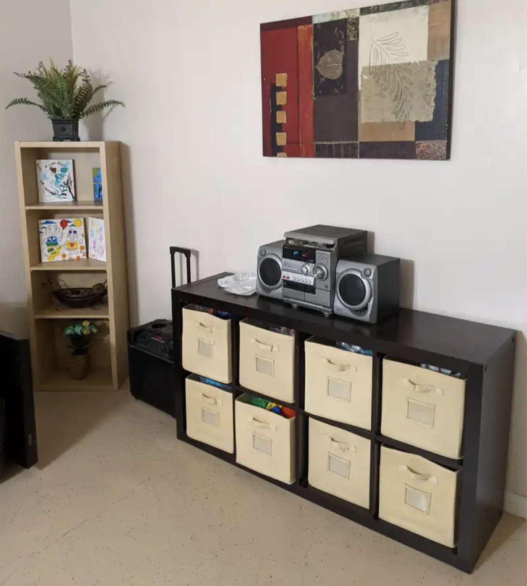 Living room corner with a tall wooden bookshelf holding potted plants and artwork, a black storage cubby with fabric bins, a black table with a stereo system, and an abstract wall art piece.
