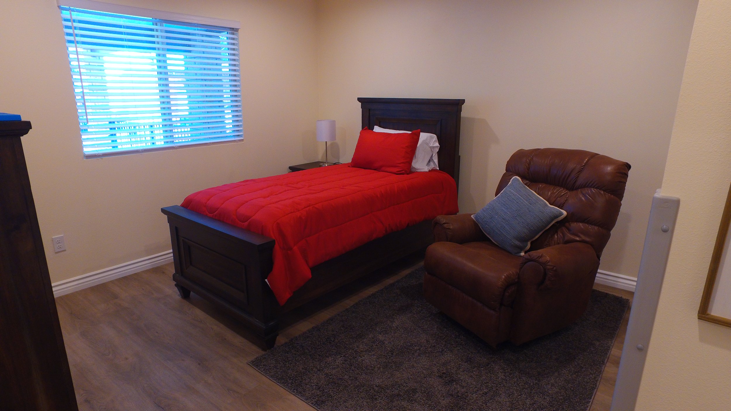 A bedroom with a twin bed covered in a red quilt, a matching pillow, white sheets, and a dark wooden headboard. Next to the bed is a small nightstand with a lamp. There is a brown leather armchair with a blue pillow on a rug, near a wall with a windo