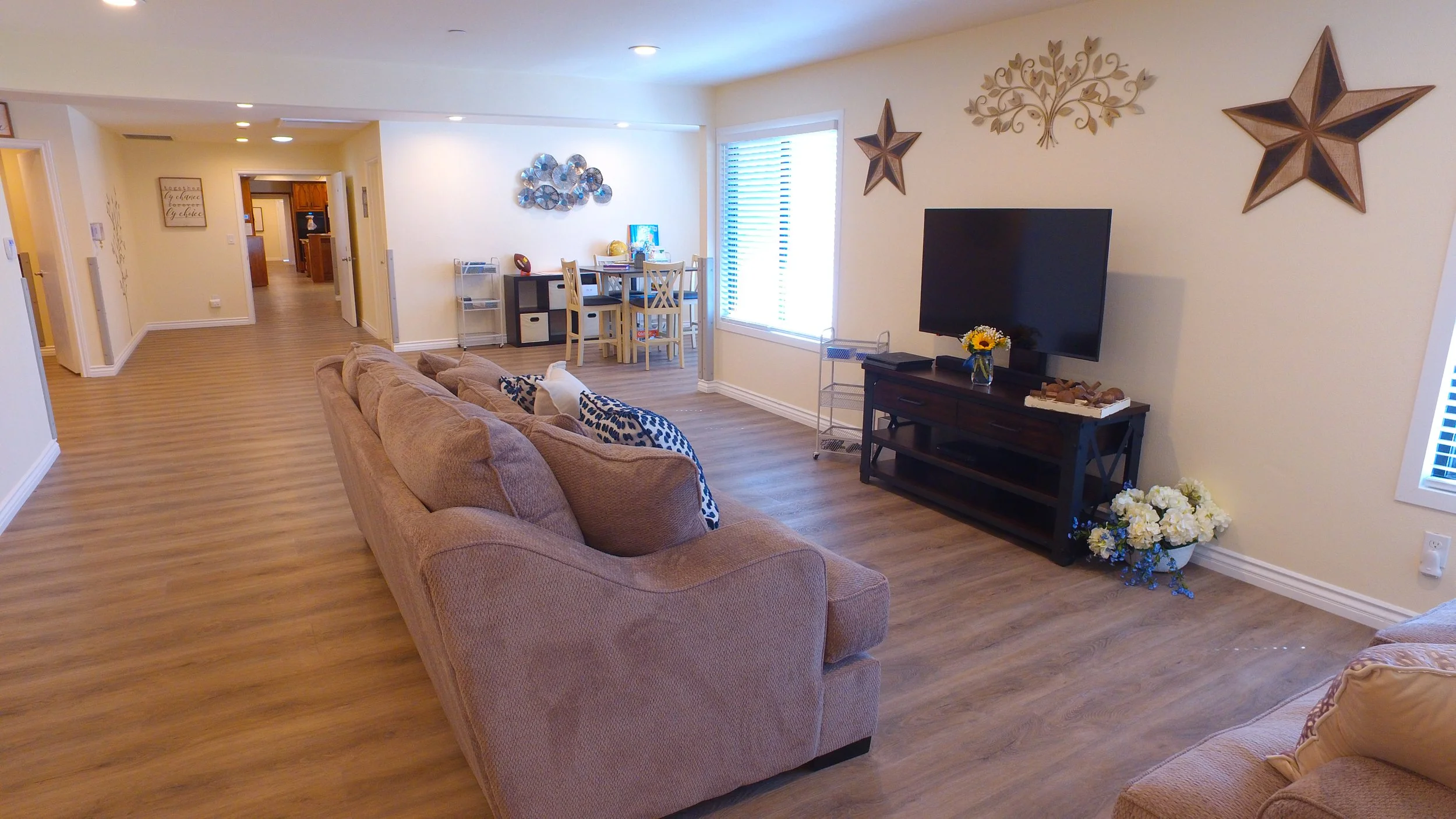Living room with beige sofas, a black TV stand with a flat-screen TV, wall decorations including stars and a tree, a dining table with chairs, and wooden flooring.