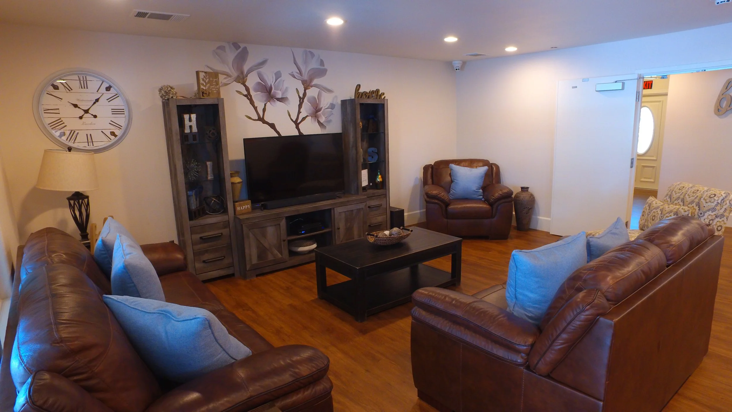 Living room with brown leather sofas, blue pillows, a black coffee table, a wooden TV stand with a television, and a wall clock. There is a decorative wall art of flowers above the TV, and various decorative items on the furniture.