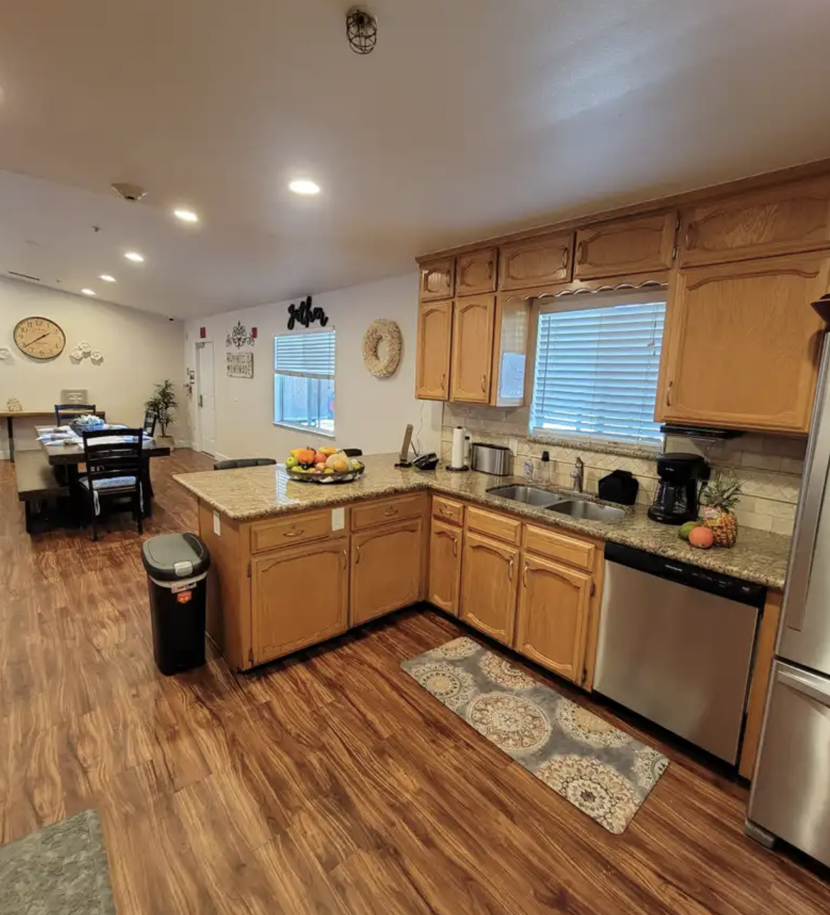 Kitchen with wooden cabinets, granite countertops, a double sink, and a window with blinds. There is a fruit bowl on the counter, along with a coffee maker, pineapple, and pumpkins. A patterned rug is on the floor, and a black trash can is nearby.