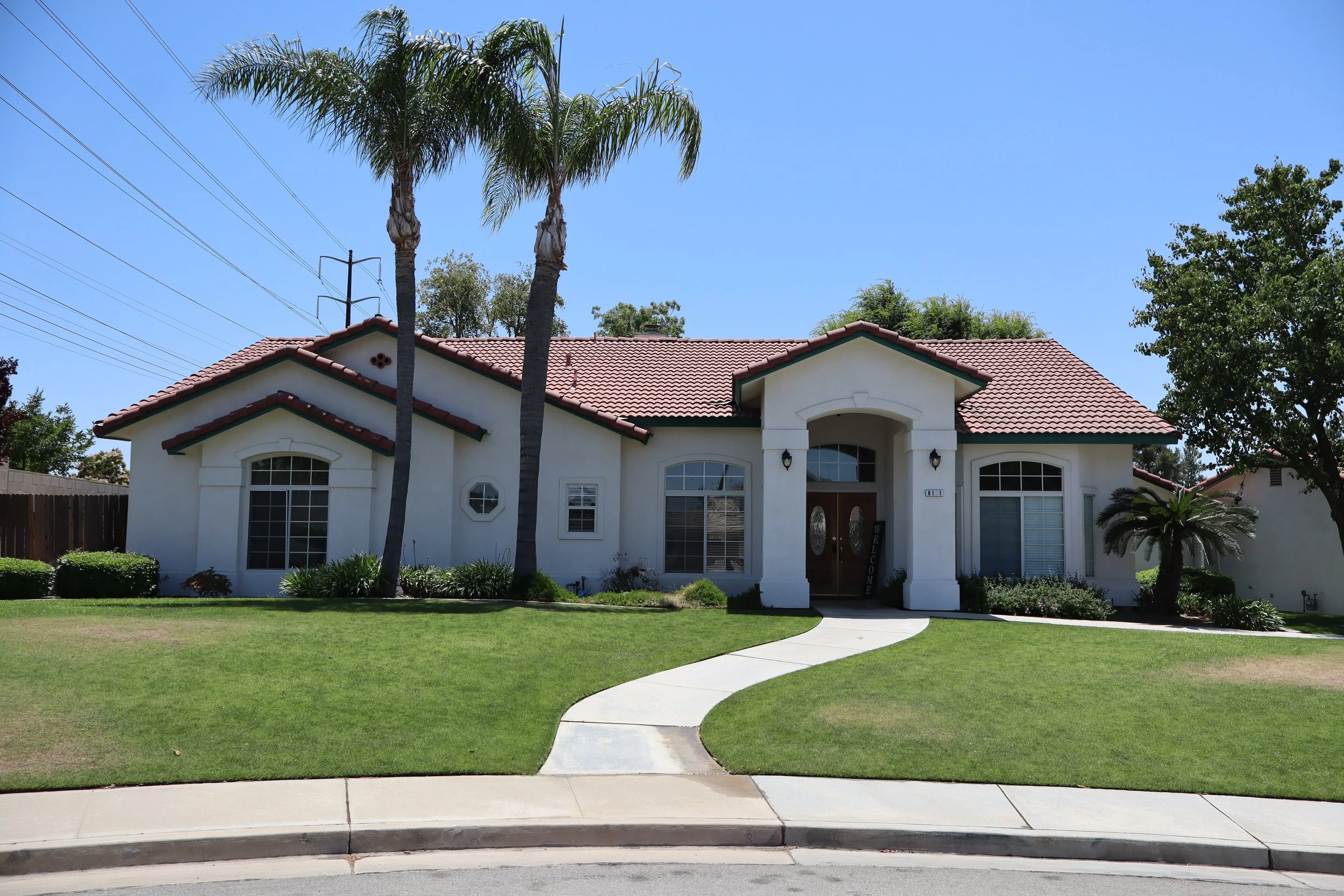 Front view of a white house with a red-tiled roof, large windows with white shutters, a curved pathway leading to the front door, palm trees, and landscaped lawn.