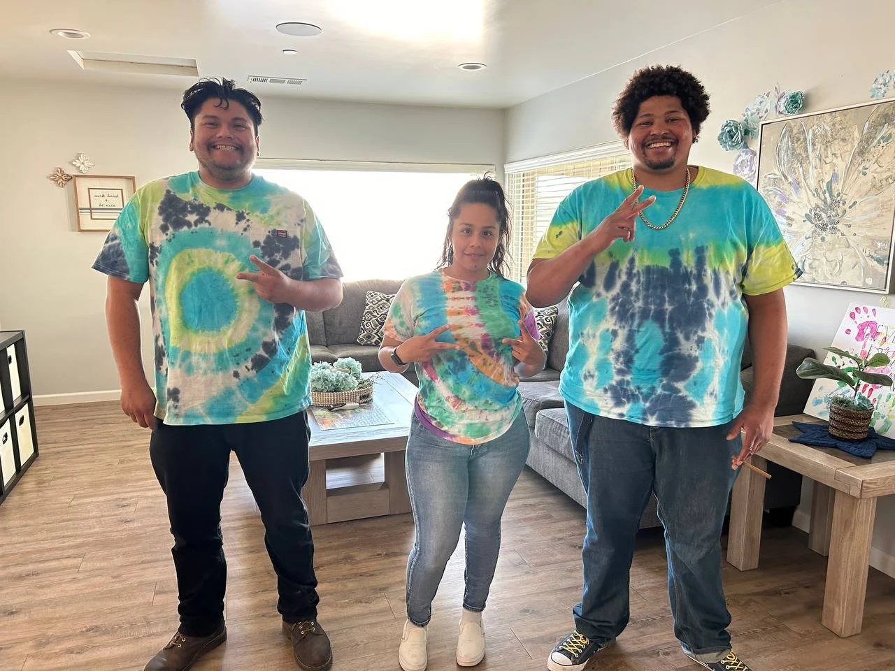 Three people wearing colorful tie-dye shirts posing in a living room. The two men on the sides are making peace signs, and the woman in the middle is holding a peace sign with both hands. The room has a beige couch, a wooden table, and floral artwork