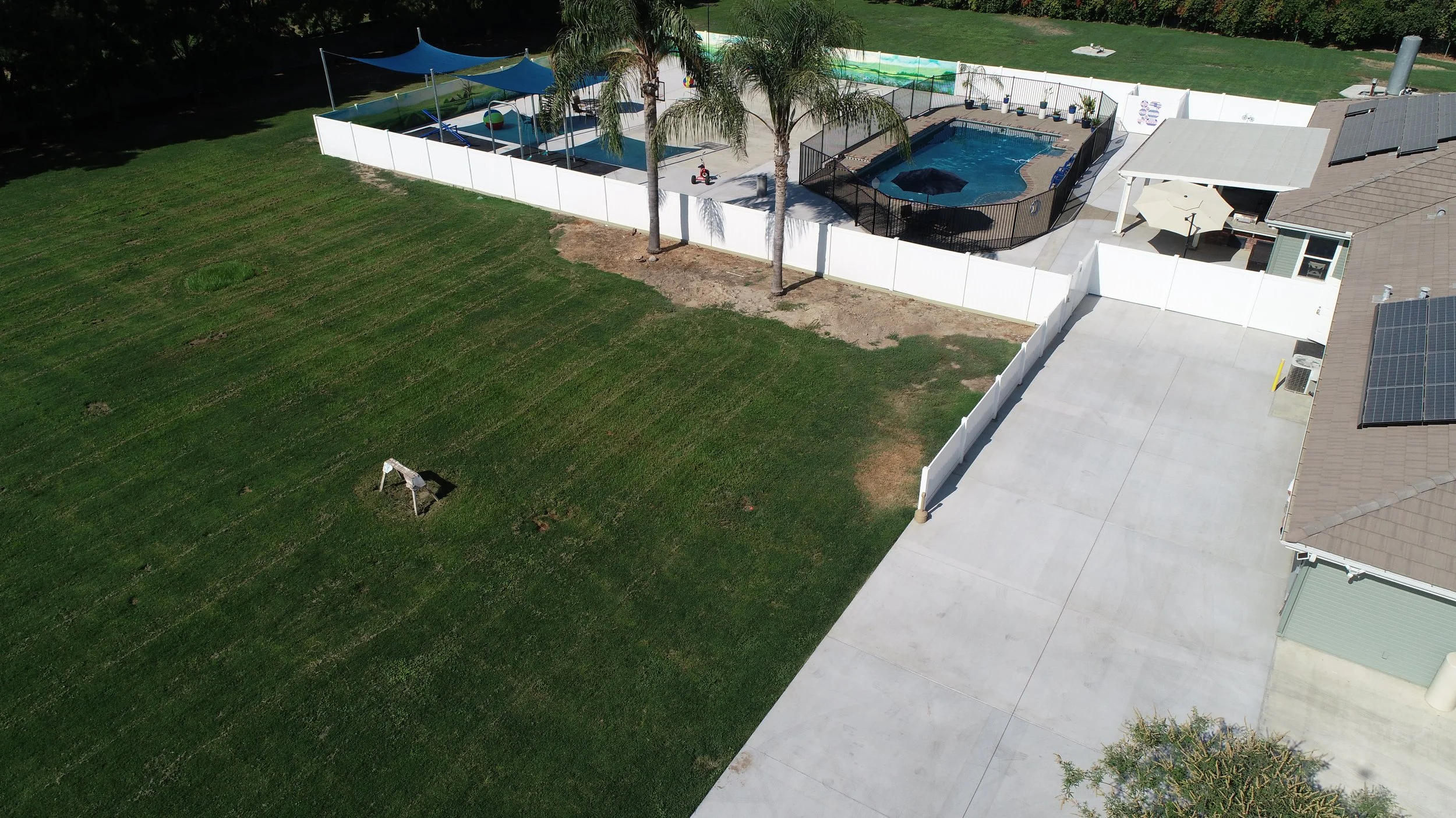 Aerial view of a backyard with a grass lawn, a white fence enclosing a pool area with a hot tub, palm trees, shaded seating area, and a large concrete driveway leading to the house.