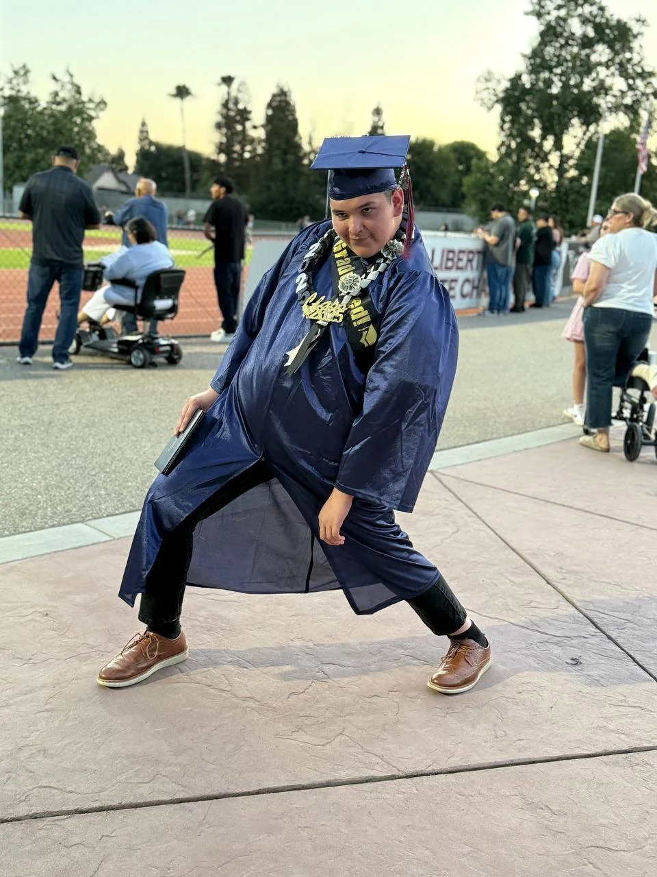 A young man in a graduation gown and cap striking a playful pose outdoors with people gathered behind him, some standing and some seated in wheelchairs, at a graduation ceremony.