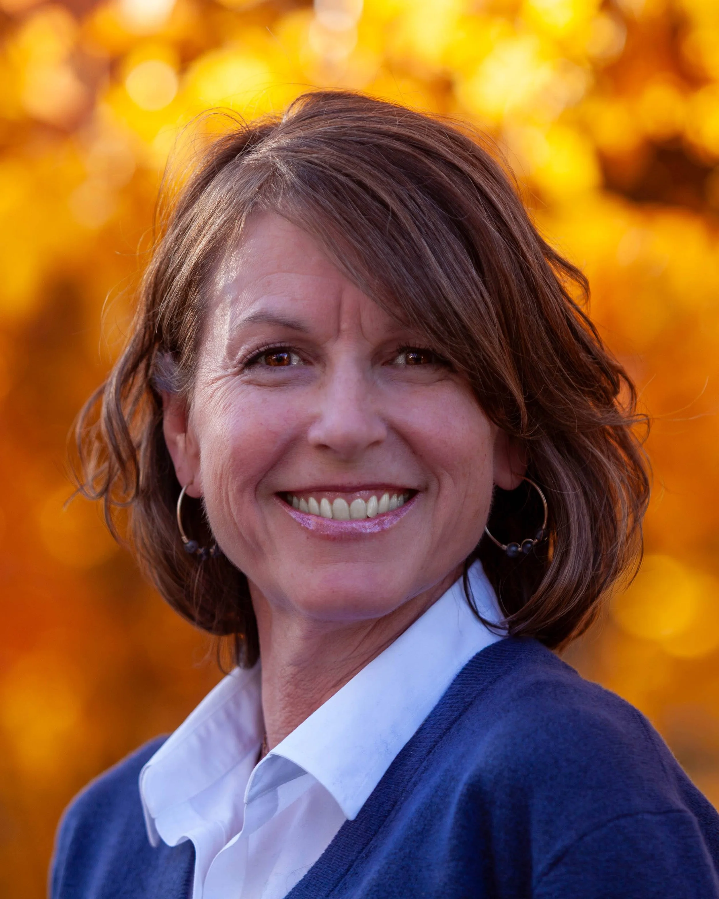 Smiling woman with short brown hair and hoop earrings, wearing a white shirt and navy sweater, outdoors with golden autumn leaves in the background.