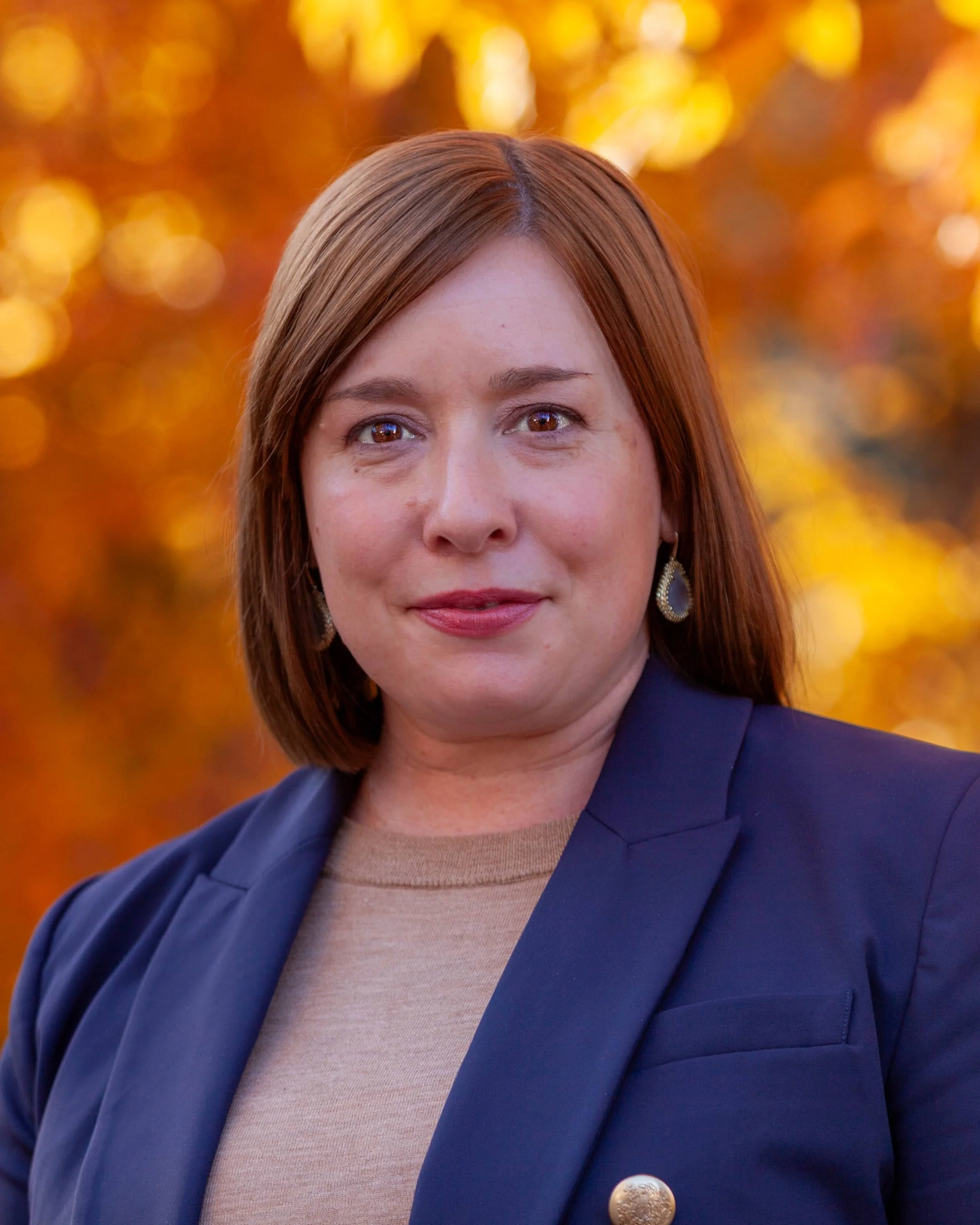 A woman with shoulder-length reddish-brown hair, wearing earrings, a beige top, and a navy blazer, standing outdoors with autumn-colored foliage in the background.