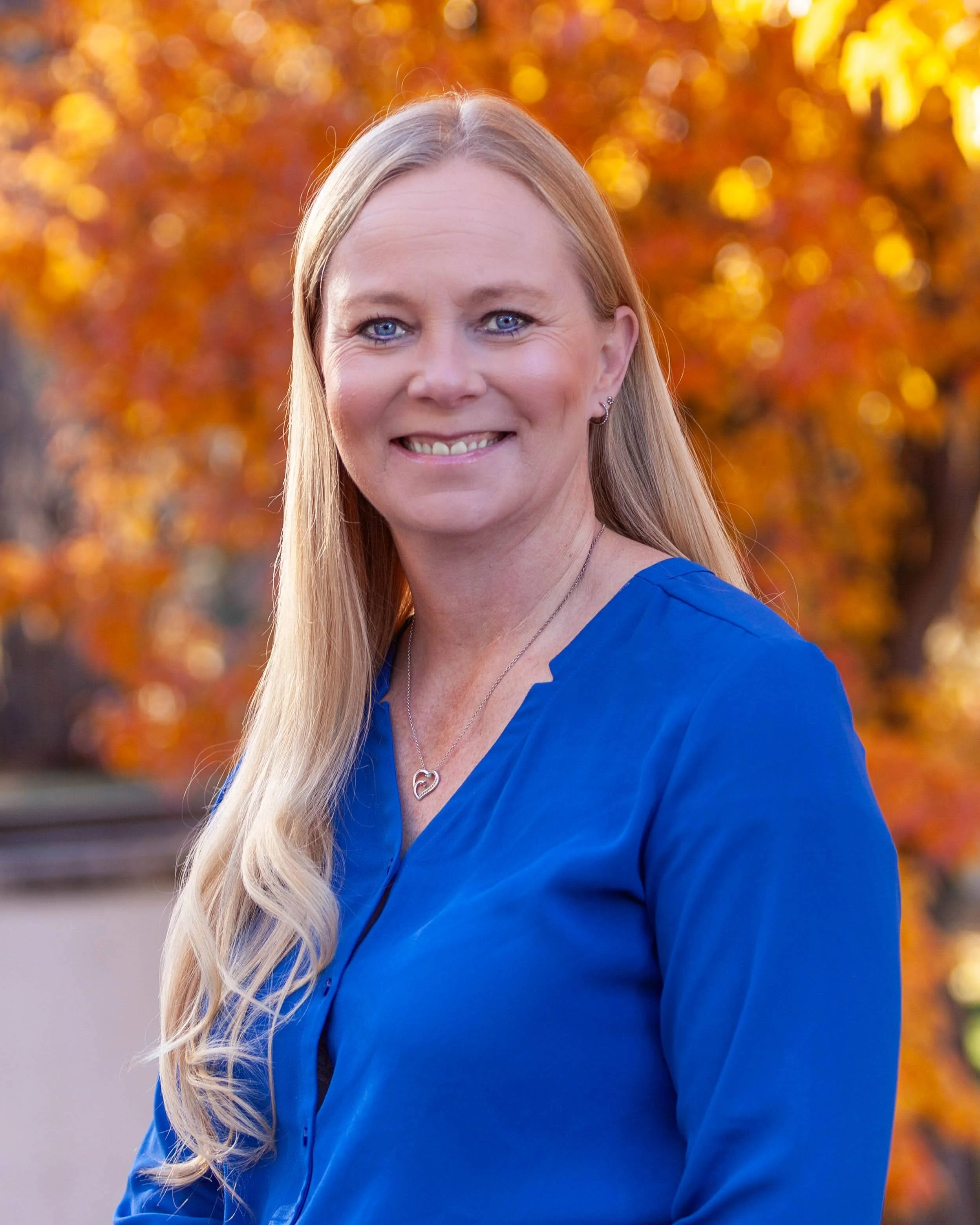 A smiling woman with long blonde hair and blue eyes wearing a blue blouse and jewelry, standing outdoors with autumn foliage in the background.