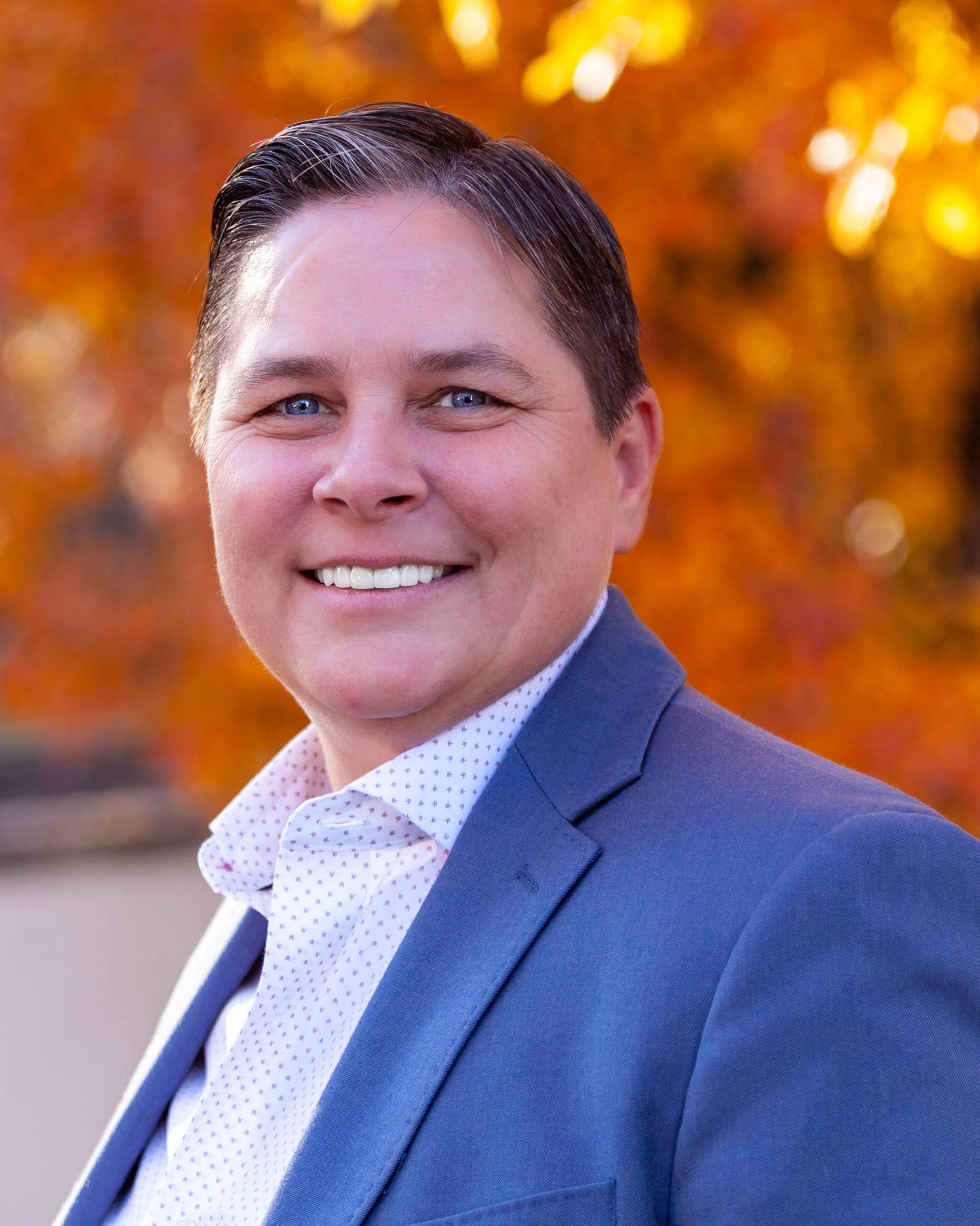 A woman with short dark hair, blue eyes, and a broad smile, wearing a blue suit jacket and white shirt with small blue patterns, standing outdoors with a blurred background of autumn orange and yellow leaves.